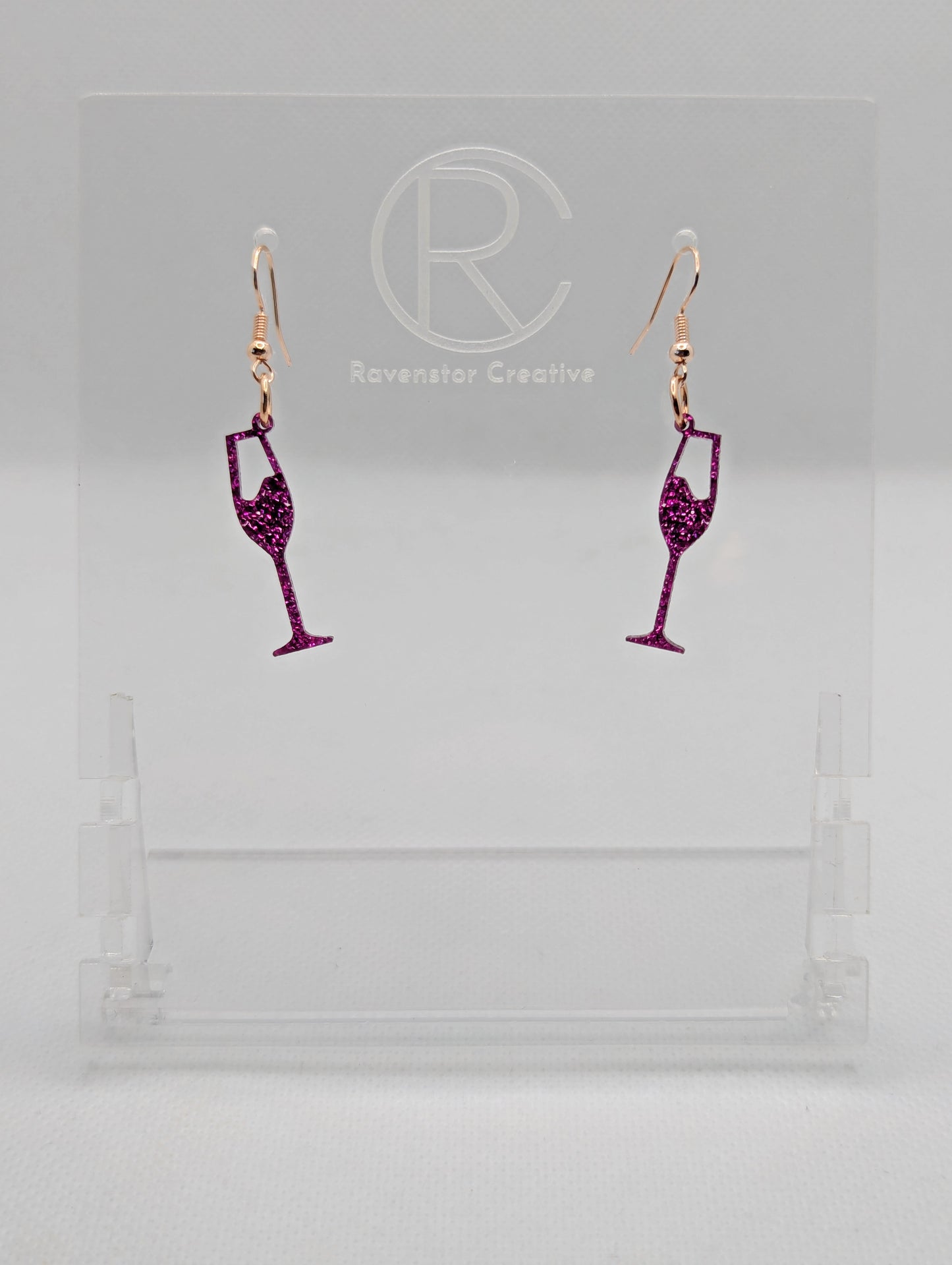 An image of a pair of Champaign Glass Earrings with Rose Gold ear wire hook fixings. These are ideal for a Hen Do or Bachelorette Party. The colour is Pink Glitter. They are on clear stands against a white background.