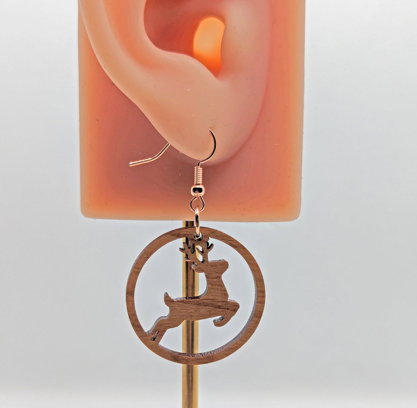 Image of a pair of circle earrings a prancing deer/reindeer cut out, on a hand showing the size against a white background. They are cherry wood and have rose gold fixings.
