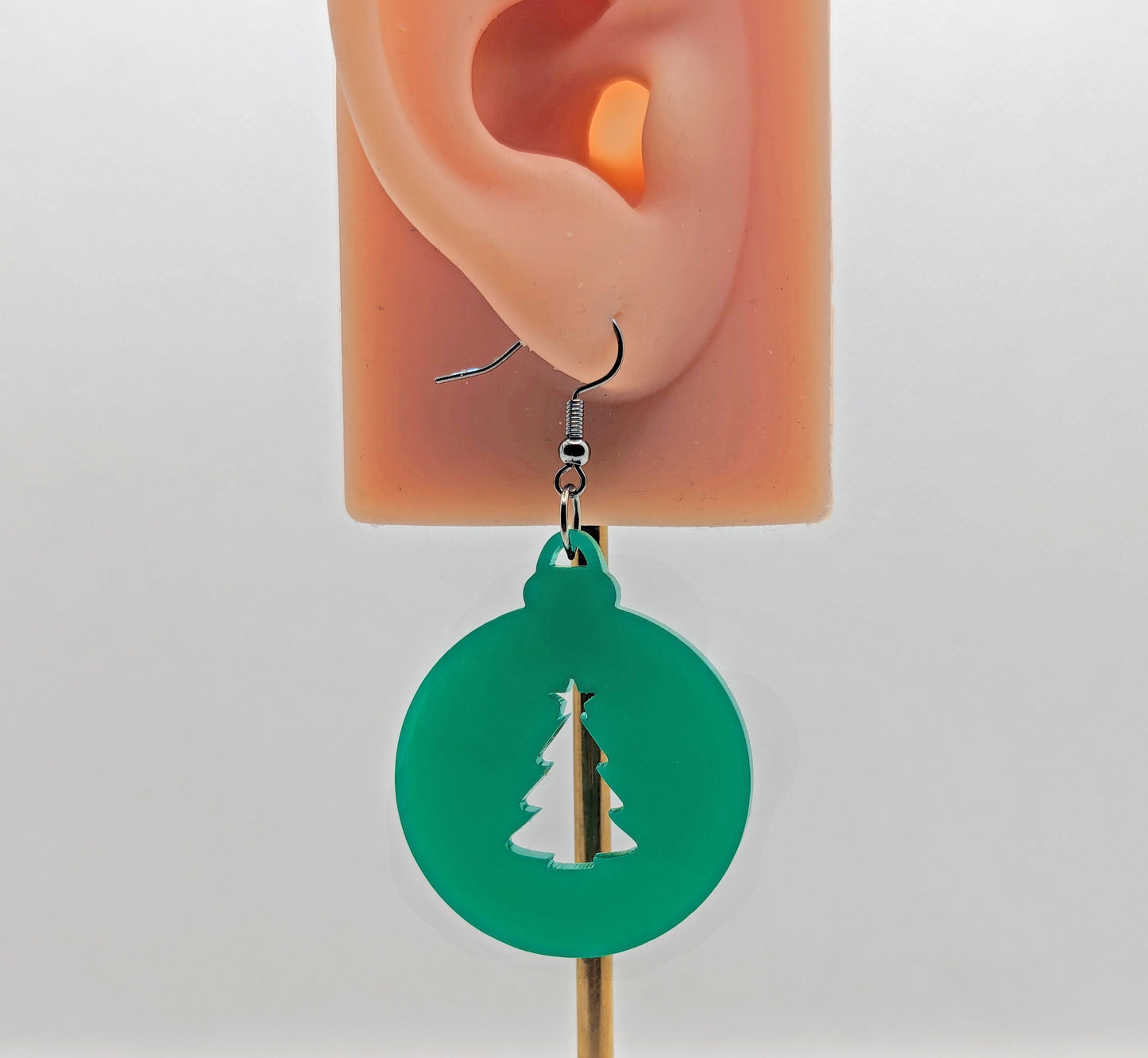 Image of a pair of earrings. They are Christmas bauble shapes with an Xmas tree cut out in Green on an ear showing the size against a white background. They and have silver fixings.