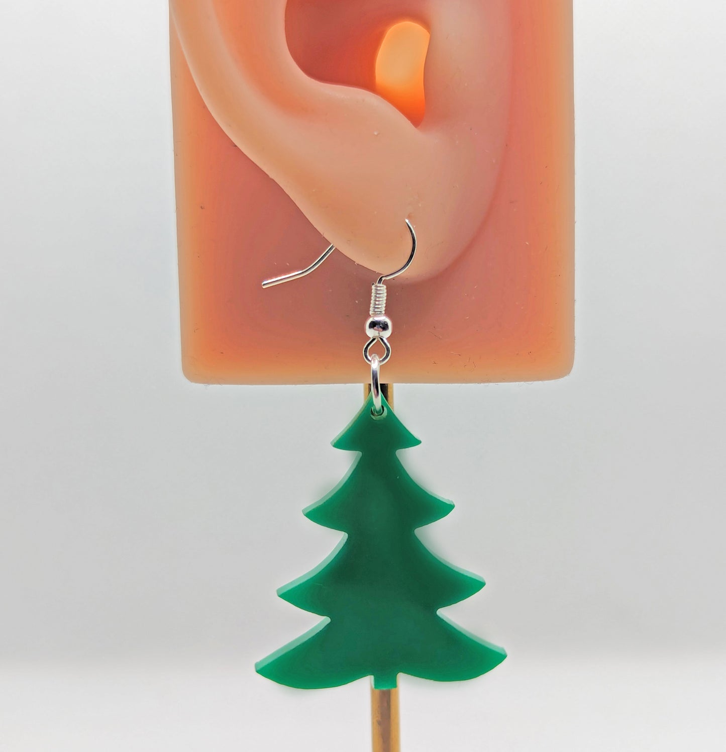 An Image showing a pair of Christmas Tree earrings in Green with silver fixings. They are against an ear to show the size. They are against a white background.