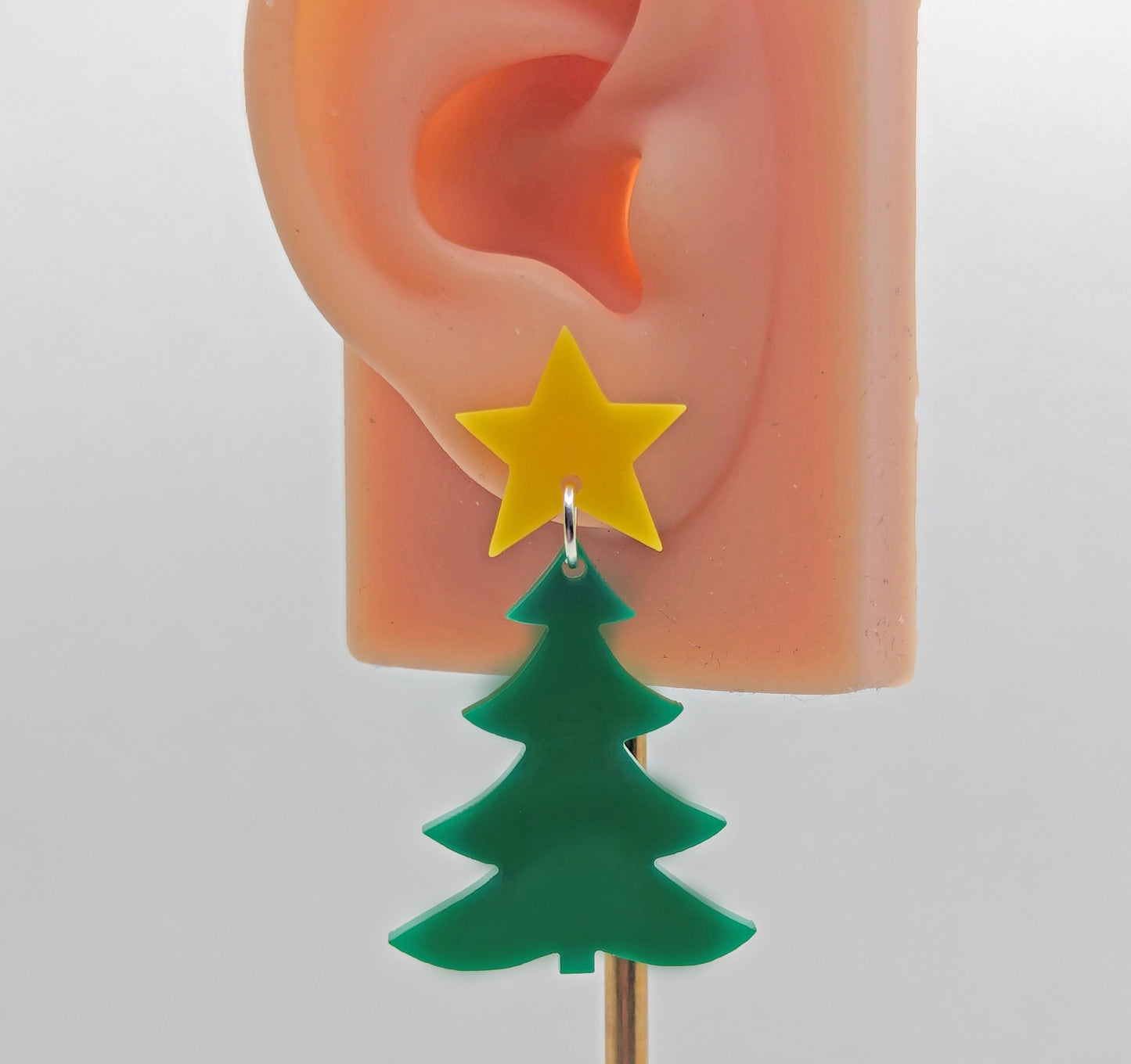 A pair of Christmas tree earrings in two parts that have stud fixings. The stud are attached to a star with the tree dangling under. They are against an ear to show the size. They are yellow for the star and green for the tree