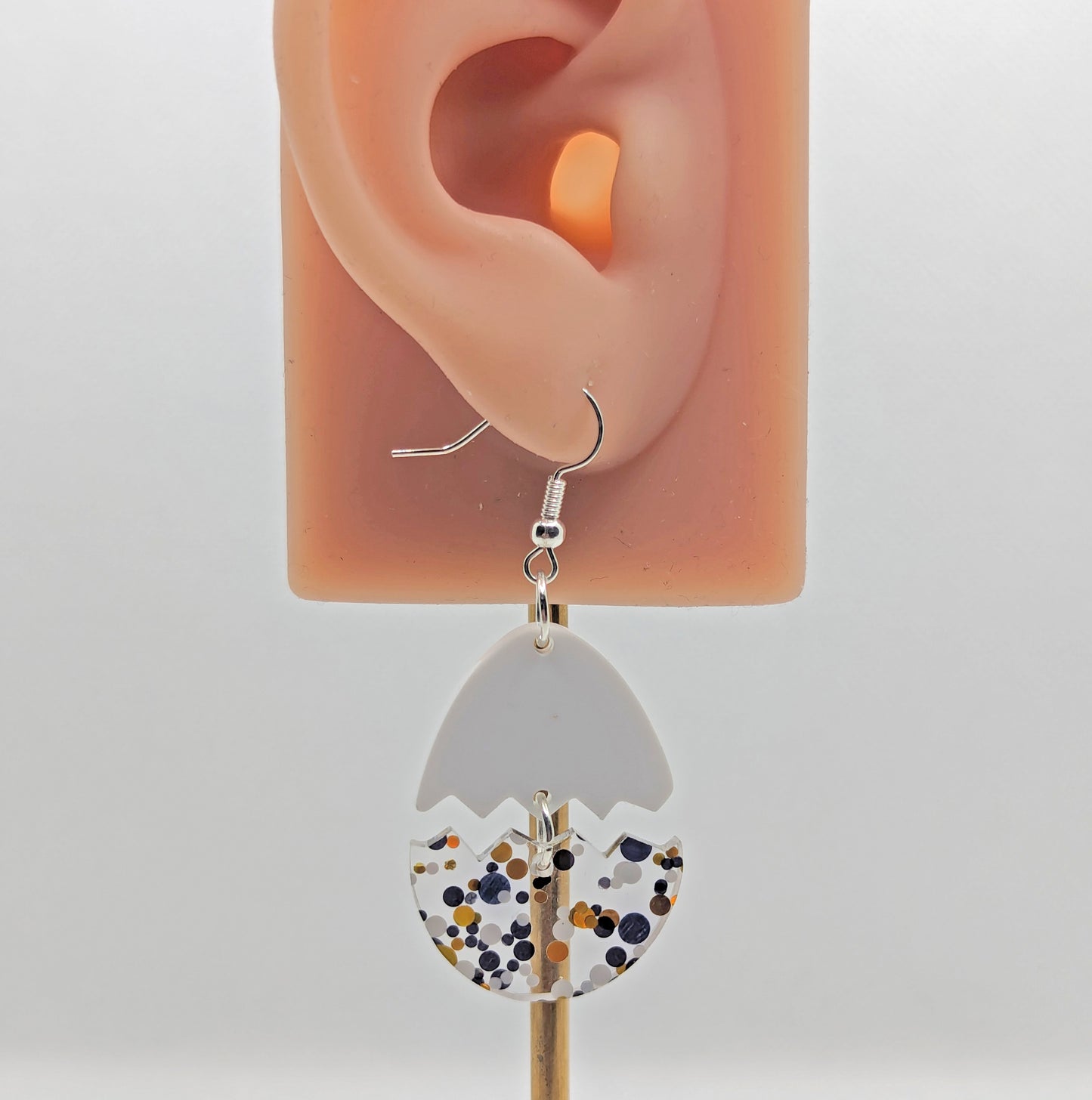 A pair of Easter egg earrings cracked in the middle separating it into two parts. They are against an ear showing the size. They have ear wire hook fixings that attach to the top which is white and the bottom dangles under, which is lollipop glitter.