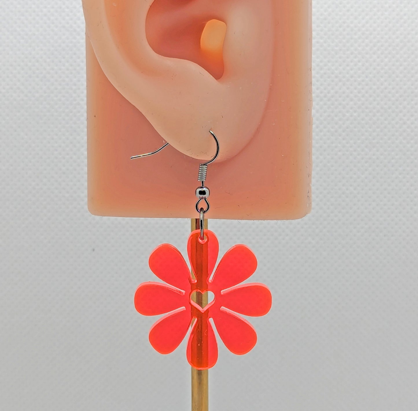 Shows an image of a Daisy flower earring with a heart cut out the middle. It is on an ear showing the size against a white background. It is neon pink and have black fixings.