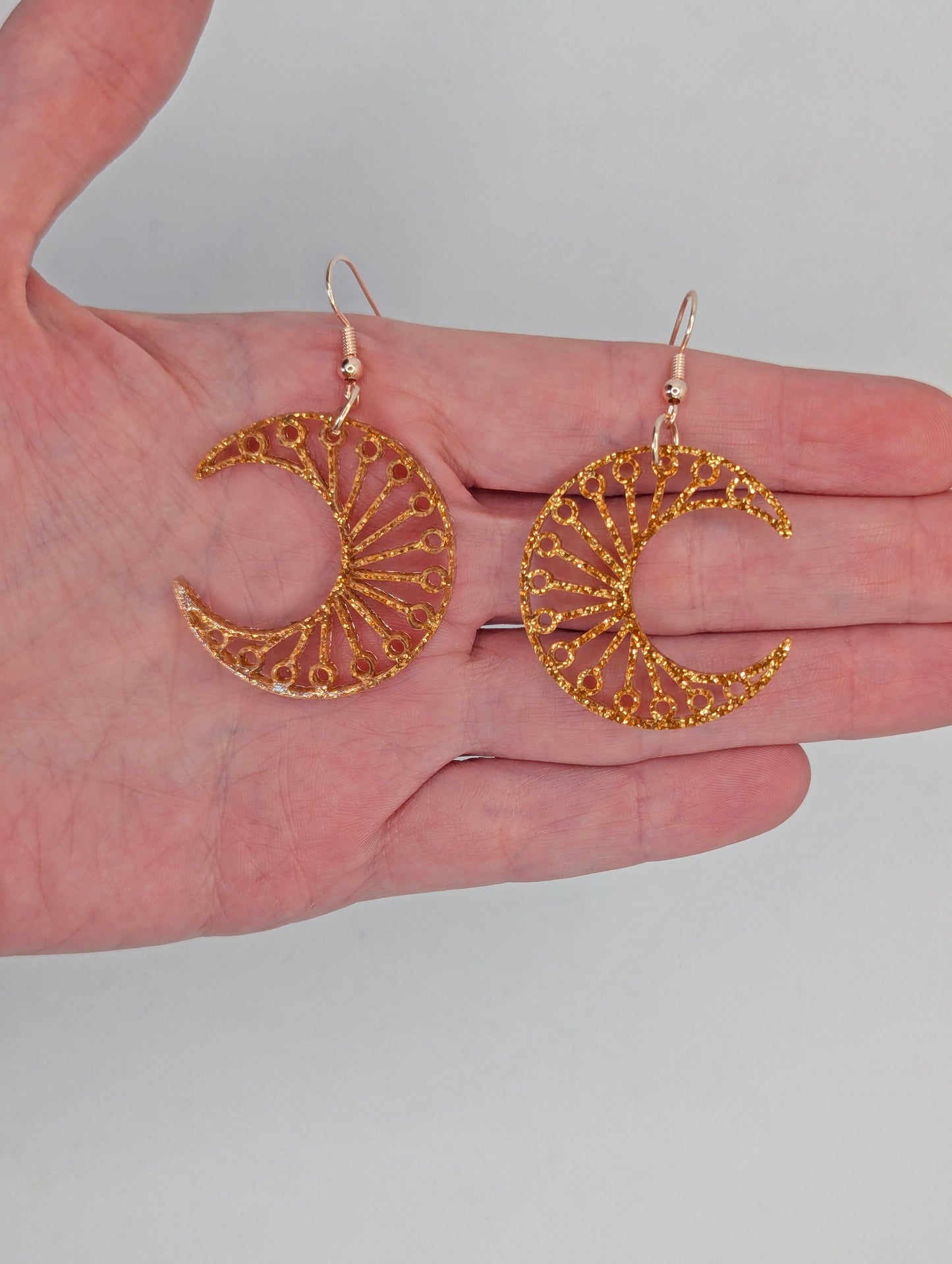 Shows a pair of earrings that have Rose Gold ear wire hook fixings. They are Decorative Crescent Moon earrings. The colour of them is Copper Glitter. They are against a white background against a hand to show the size of two finger widths in length and width.