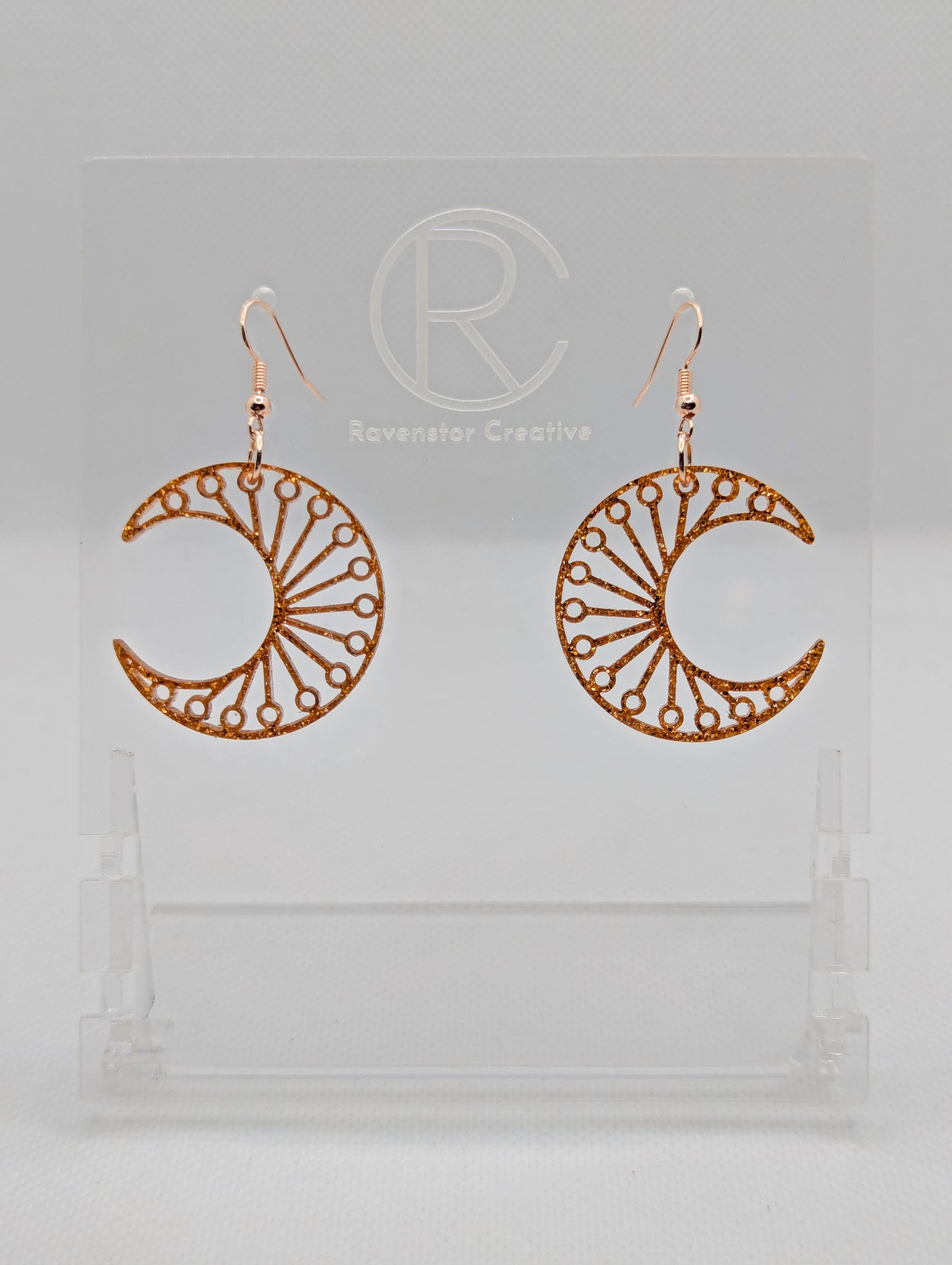 Shows a pair of earrings that have Rose Gold ear wire hook fixings. They are Decorative Crescent Moon earrings. The colour of them is Copper Glitter. They are against a white background on see through stands with the Ravenstor Creative logo.