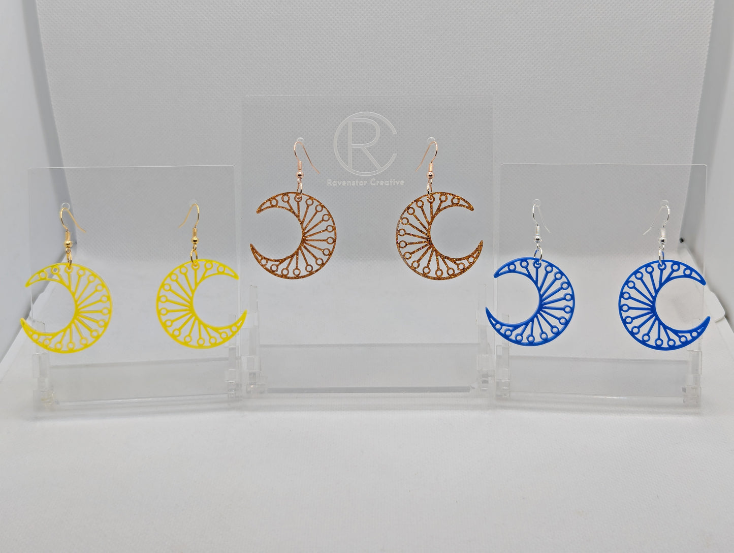 Shows three pairs of earrings that have ear wire hook fixings. They are Decorative Crescent Moon earrings. The colours are Neon Orange, copper glitter and Blue. They are against a white background on see through stands with the Ravenstor Creative logo.