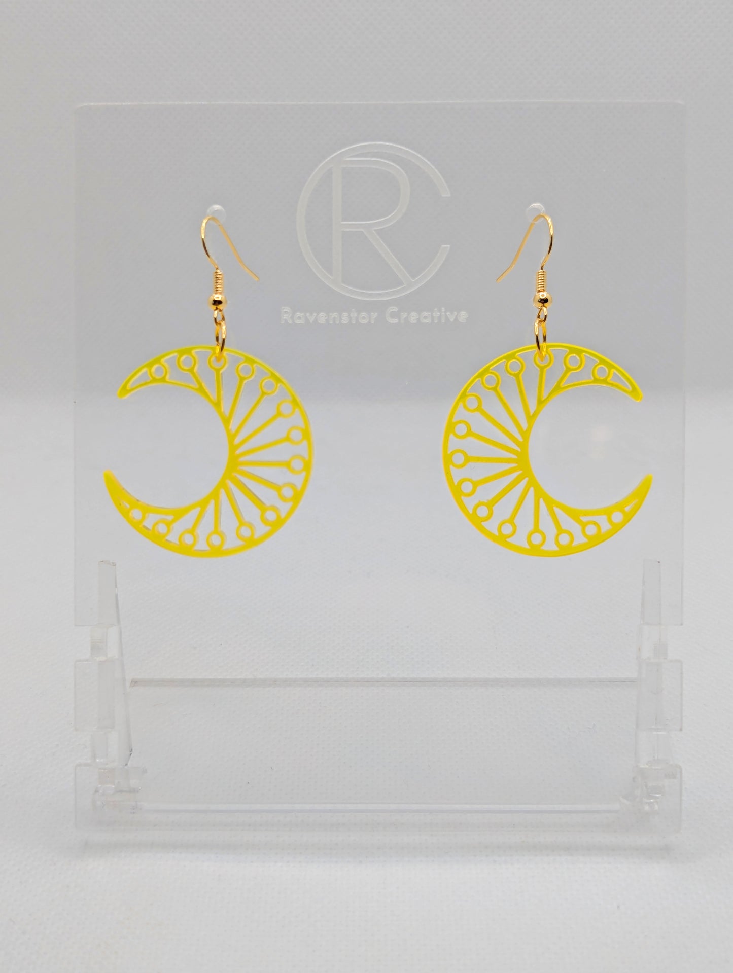 Shows a pair of earrings that have Gold ear wire hook fixings. They are Decorative Crescent Moon earrings. The colour of them is Neon Orange. They are against a white background on see through stands with the Ravenstor Creative logo.