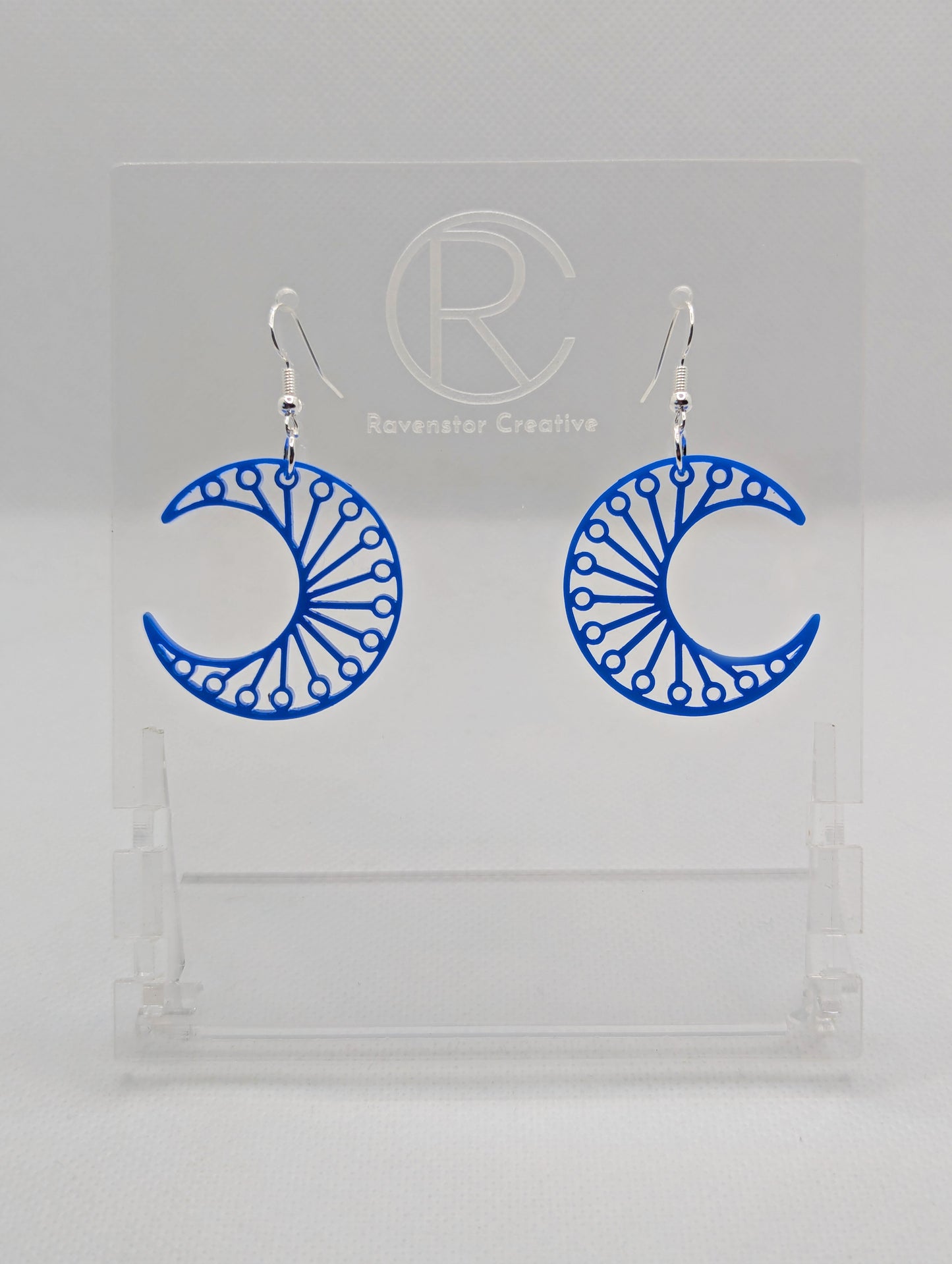Shows a pair of earrings that have Silver ear wire hook fixings. They are Decorative Crescent Moon earrings. The colour of them is Blue. They are against a white background on see through stands with the Ravenstor Creative logo.
