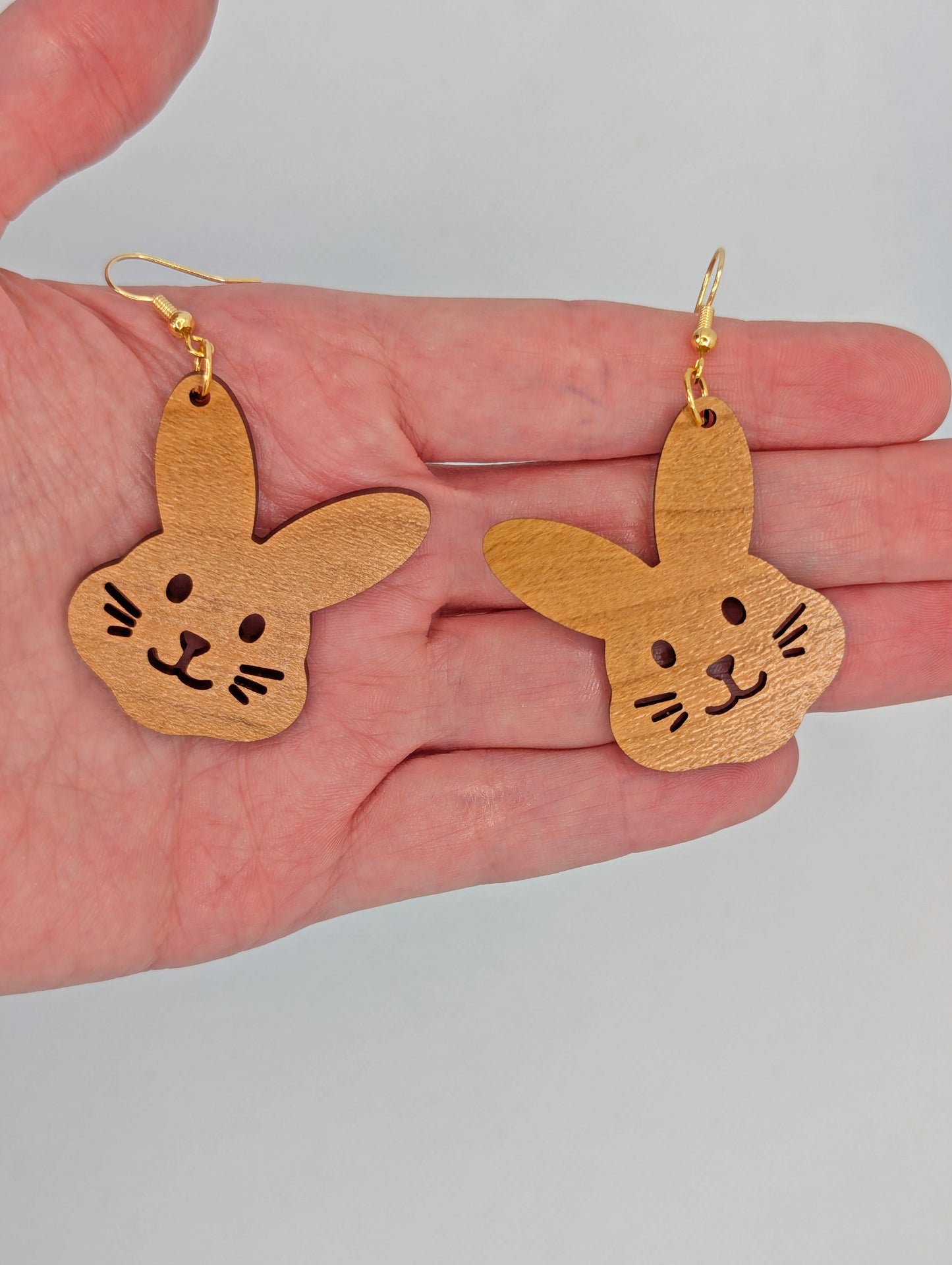 A pair of wooden earrings that have gold ear wire hook fixings. They are large bunny rabbit head shapes. They are Cherry Wood in colour and are against a white background on a hand to show the size of three finger widths in length and two and a half wide.
