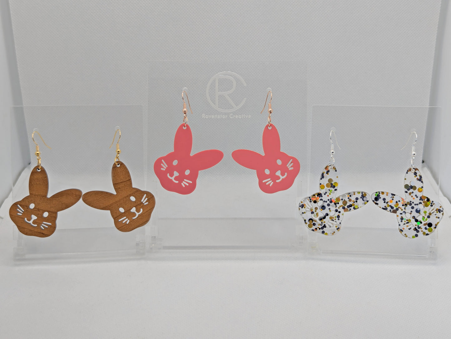 Three pairs of earrings that have ear wire hook fixings. They large bunny rabbit head earrings. The colours are Cherry Wood, Sherbet Pink and Lollipop Glitter. They are against a white background on see through stands with the Ravenstor Creative Logo on.