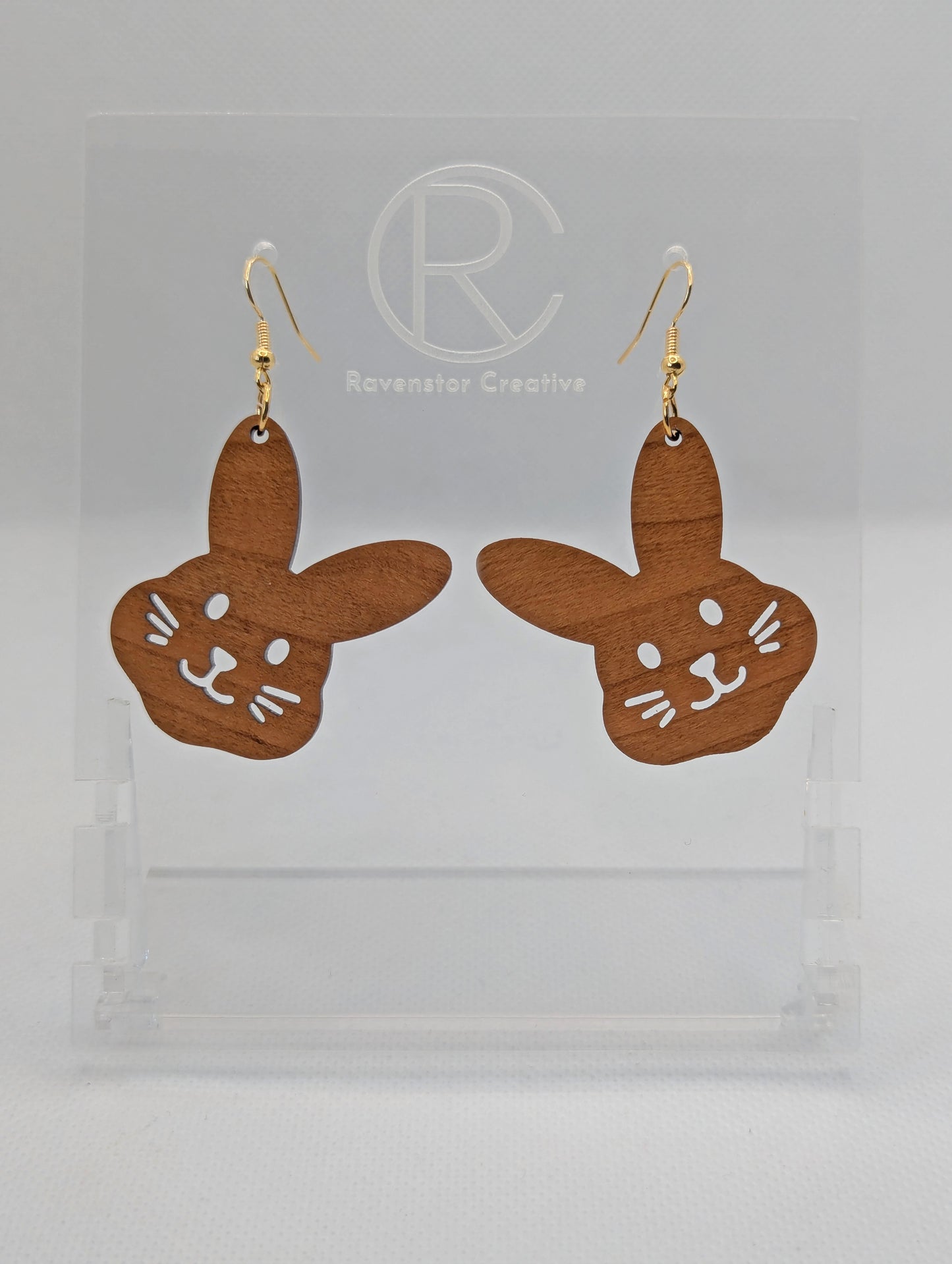 A pair of wooden earrings that have gold ear wire hook fixings. They are large bunny rabbit head shapes. They are Cherry Wood in colour and are against a white background on see through stand with the Ravenstor Creative Logo.