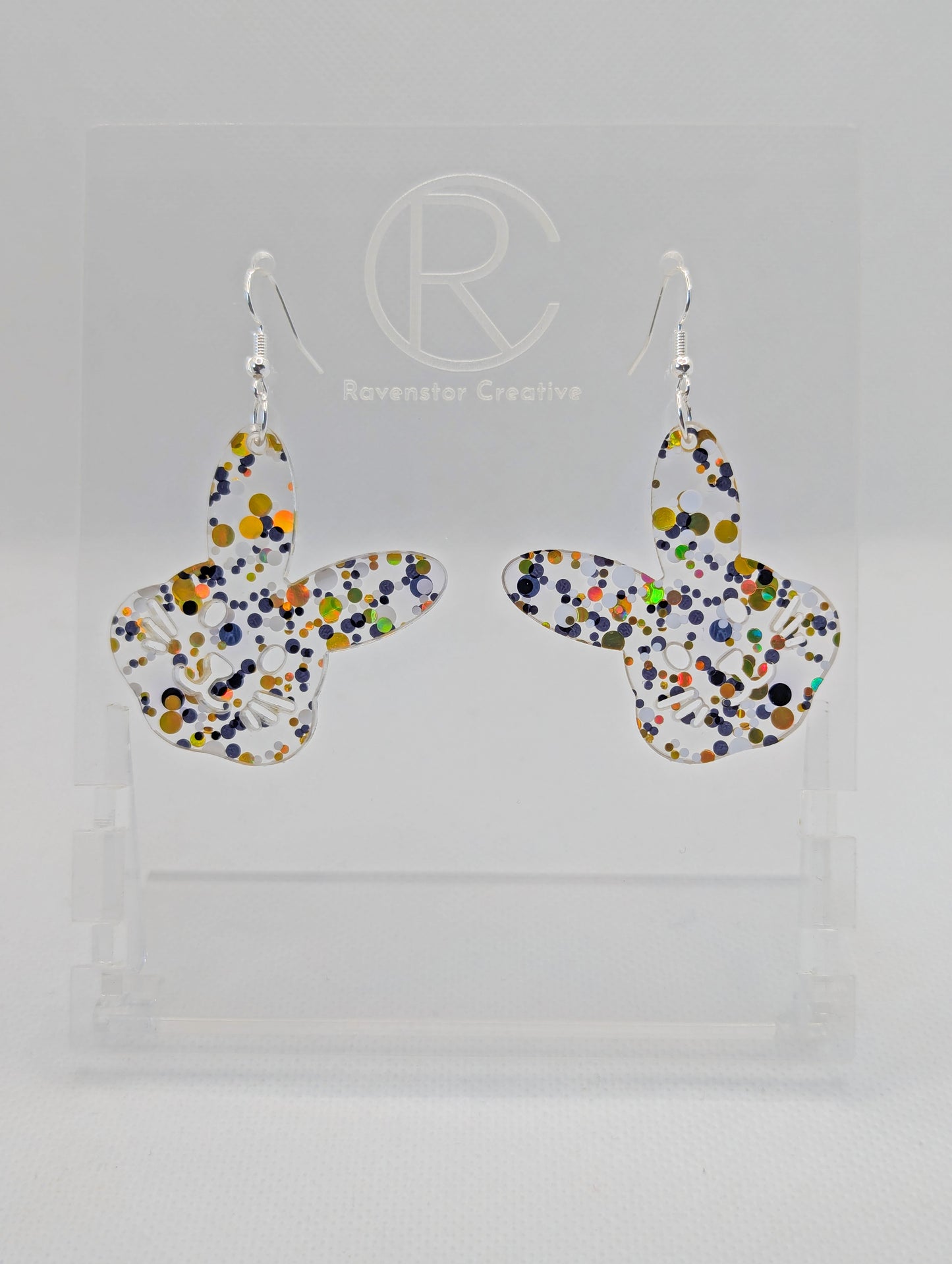A pair of wooden earrings that have silver ear wire hook fixings. They are large bunny rabbit head shapes. They are Lollipop Glitter in colour and are against a white background on see through stand with the Ravenstor Creative Logo.