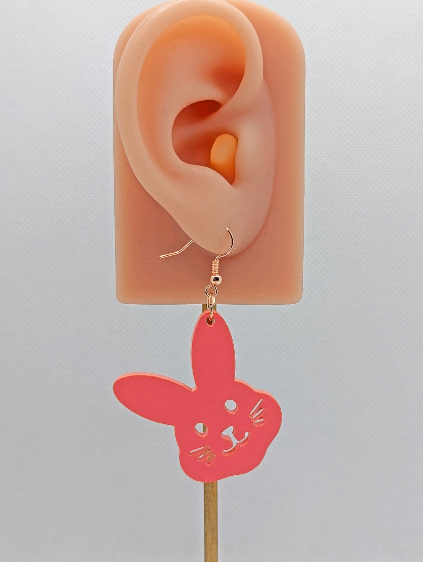 An earring that has a rose gold ear wire hook fixing. It is a bunny rabbit head shape and it cut from Sherbet Pink coloured Acrylic. They are on an ear showing the size against a white background.