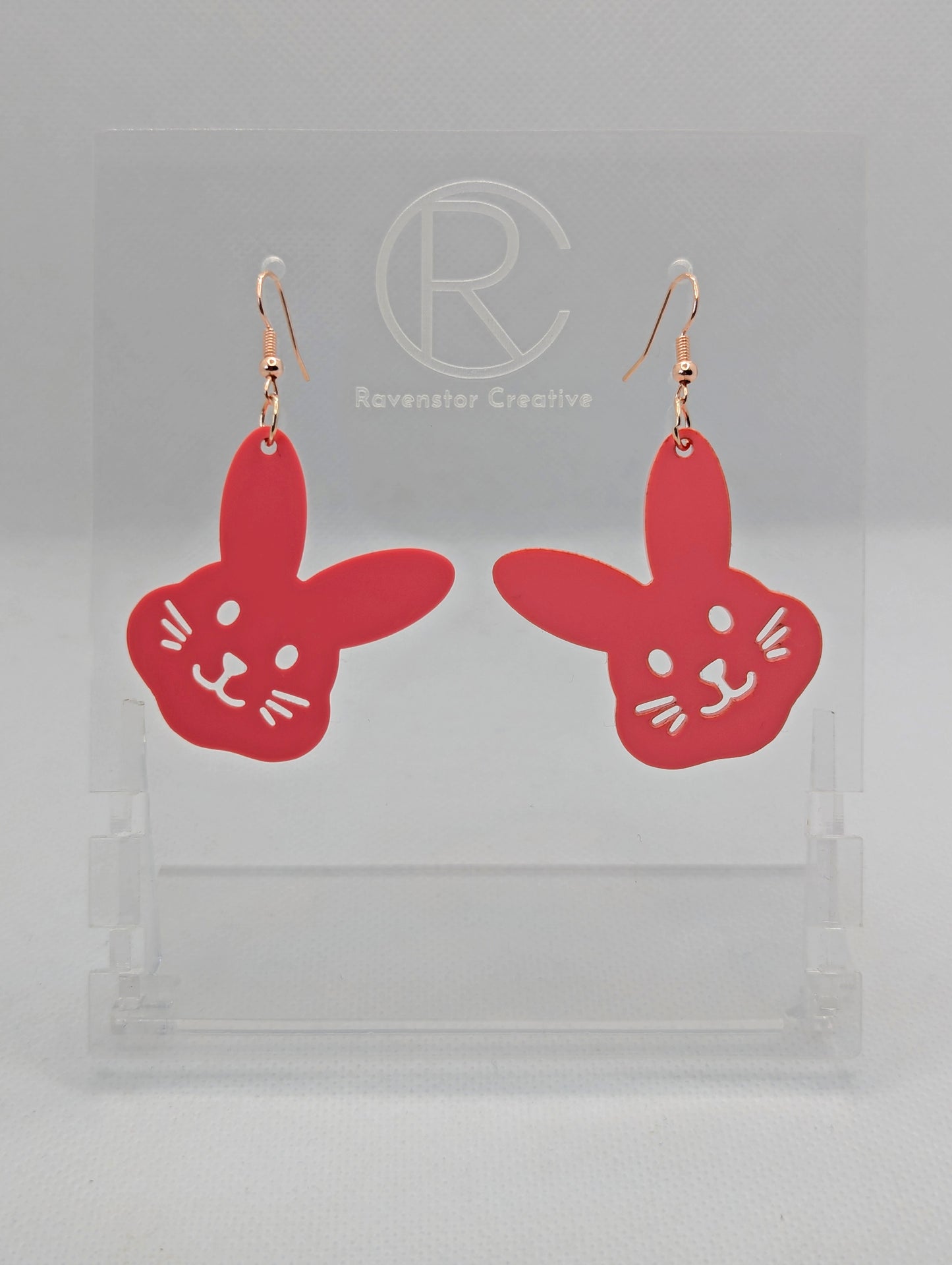 A pair of wooden earrings that have Rose Gold ear wire hook fixings. They are large bunny rabbit head shapes. They are Sherbet Pink in colour and are against a white background on see through stand with the Ravenstor Creative Logo.