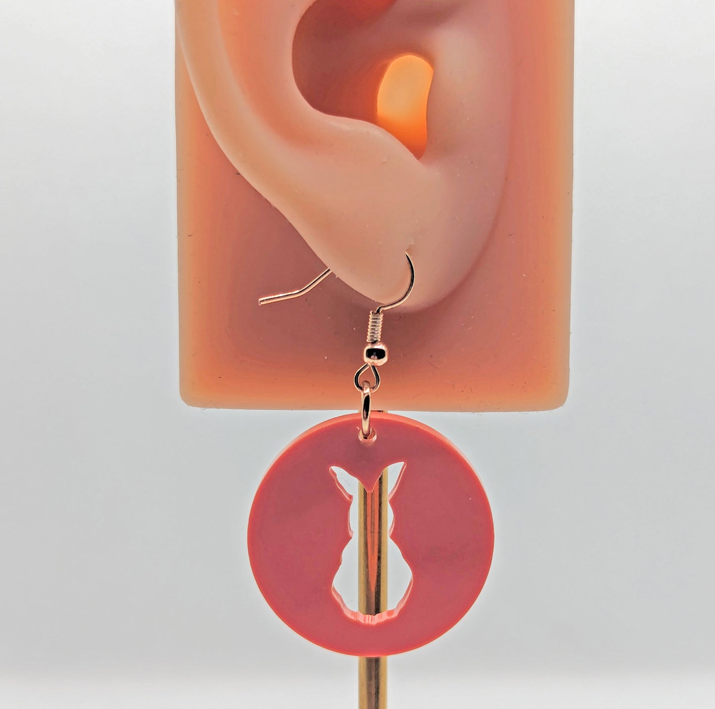 An acrylic earring that has rose gold ear wire hook fixings against an ear to show the size. it is a Circle with an Easter bunny / Rabbit cut out. It is sherbet pink in colour.