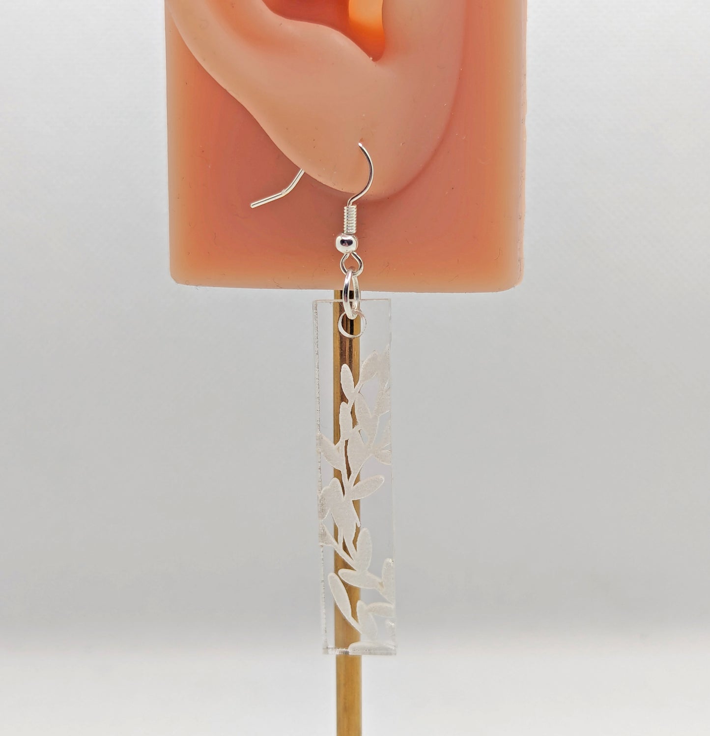 Image of a long slim rectangle earring with silver fixings on an ear showing the size. They are clear/see through and are engraved with bay leaves.