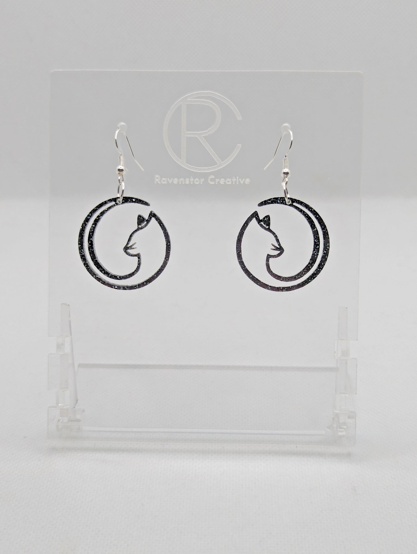 Shows a pair of acrylic earrings that have silver ear wire hook fixings. They are Full Moon inspired Cat shapes. They are in Purple Glitter and are against a white background on see through stand with the Ravenstor Creative logo on it.