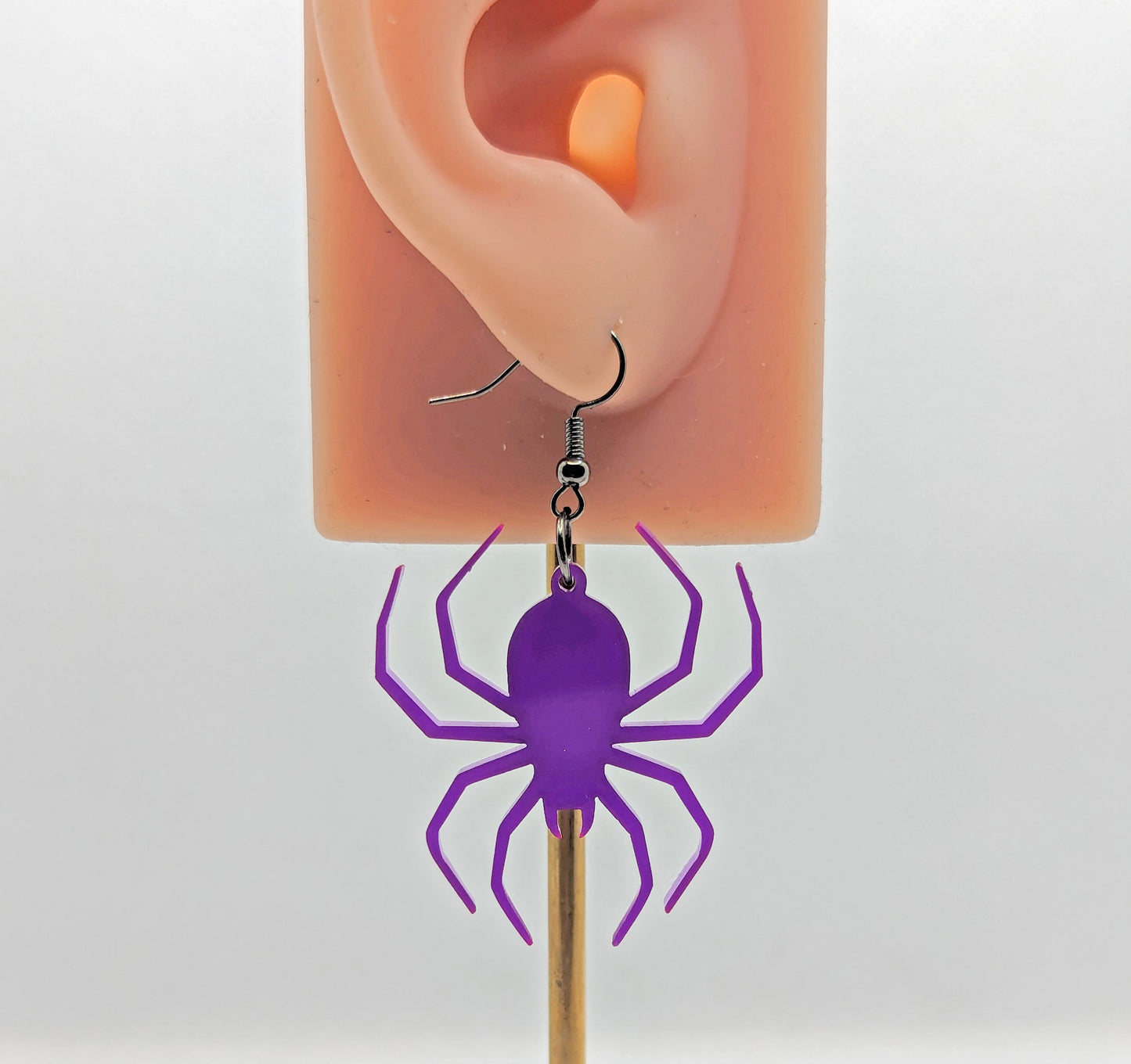 An earring on an ear to show the size with black fixings. It is a spider with thin legs like a black widow. It is purple in colour and hang like it is spinning a web. They are against a white background