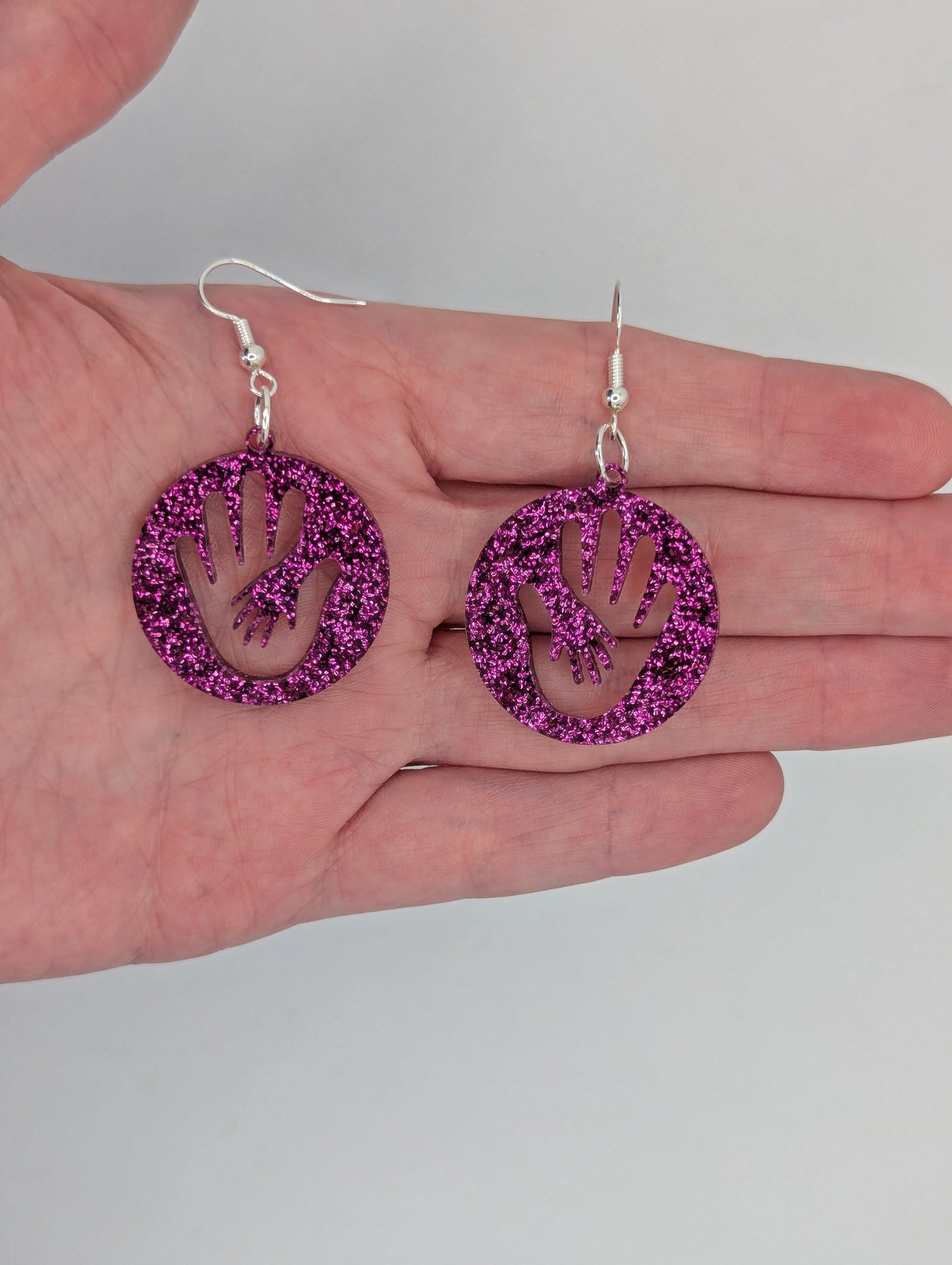 A pair of round earrings in with the image of a hand in a hand symbol cut out. They have Silver ear wire hook fixings and the colour is Pink Glitter. They are against a hand to show the size of two finger widths in length and wide.