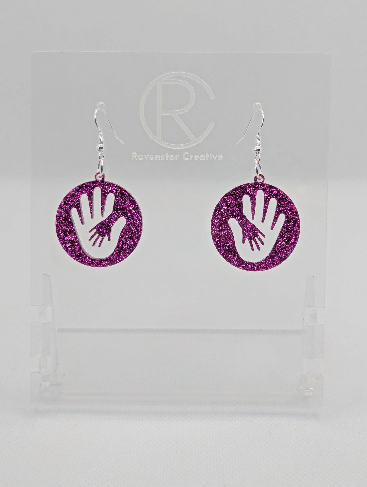A pair of round earrings in with the image of a hand in a hand symbol cut out. They have Silver ear wire hook fixings and the colour is Pink Glitter. They are on a clear stand with the Ravenstor Creative Logo on it against a white background.