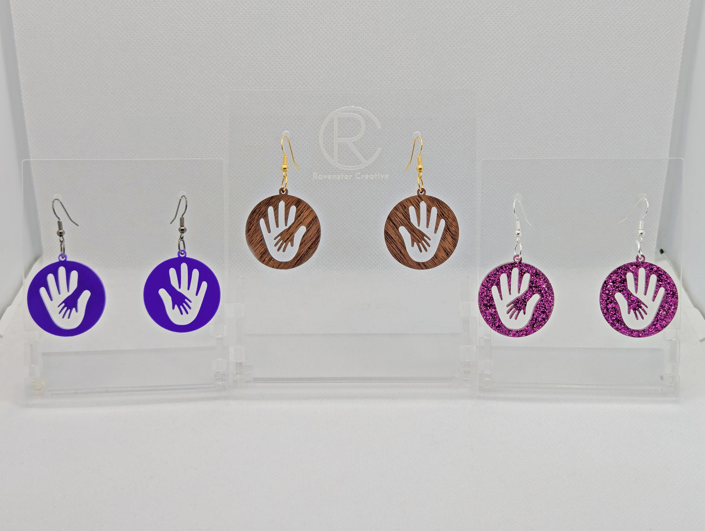 Three pairs of round earrings in with the image of a hand in a hand symbol cut out. They have ear wire hook fixings and they are Purple Acrylic, Walnut Wood and Pink Glitter Acrylic. They are on clear stands against a white background.