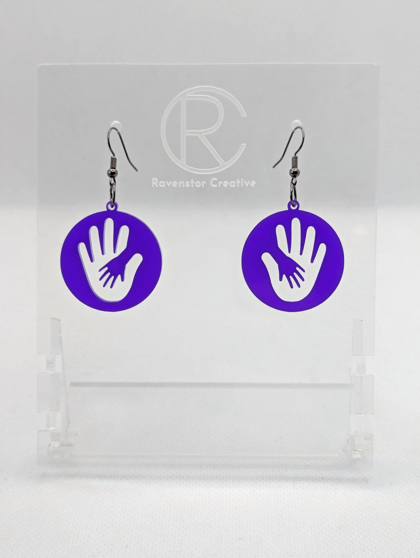 A pair of round earrings in with the image of a hand in a hand symbol cut out. They have Black ear wire hook fixings and the colour is Purple. They are on a clear stand with the Ravenstor Creative Logo on it against a white background.