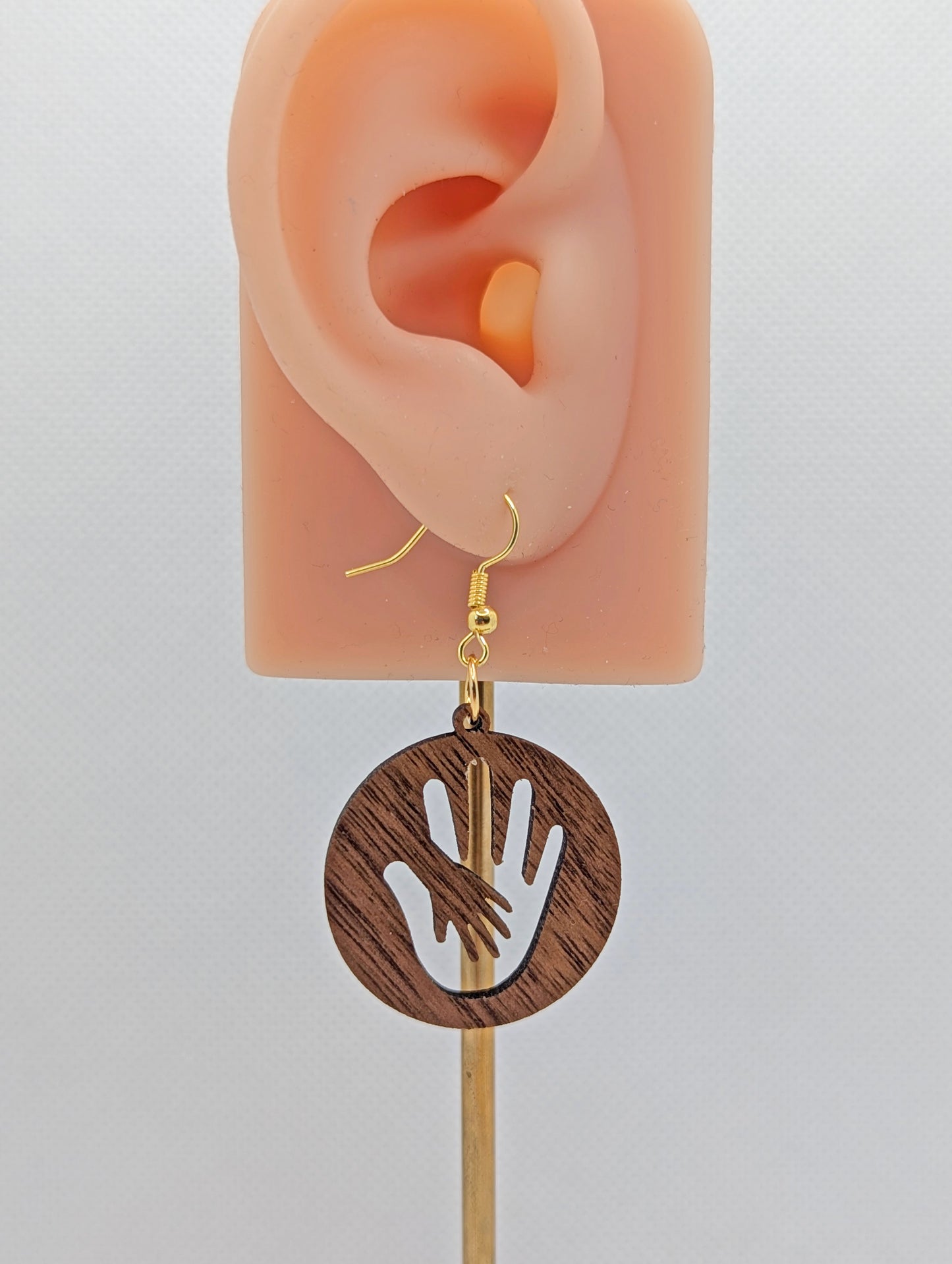 A round earring with the image of a hand in a hand symbol cut out. It has gold ear wire hook fixings and is made from Walnut wood. They are on an ear to show the size against a white background