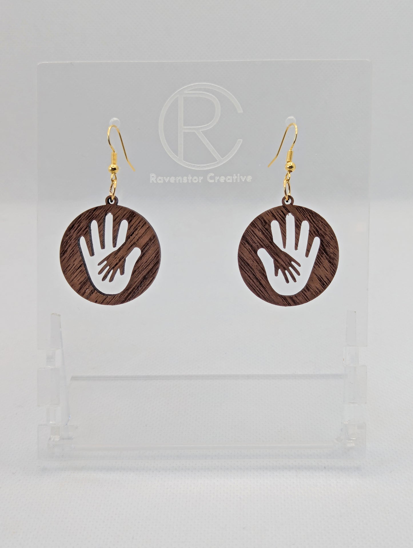A pair of round earrings in with the image of a hand in a hand symbol cut out. They have Gold ear wire hook fixings and the colour is Walnut Wood. They are on a clear stand with the Ravenstor Creative Logo on it against a white background.