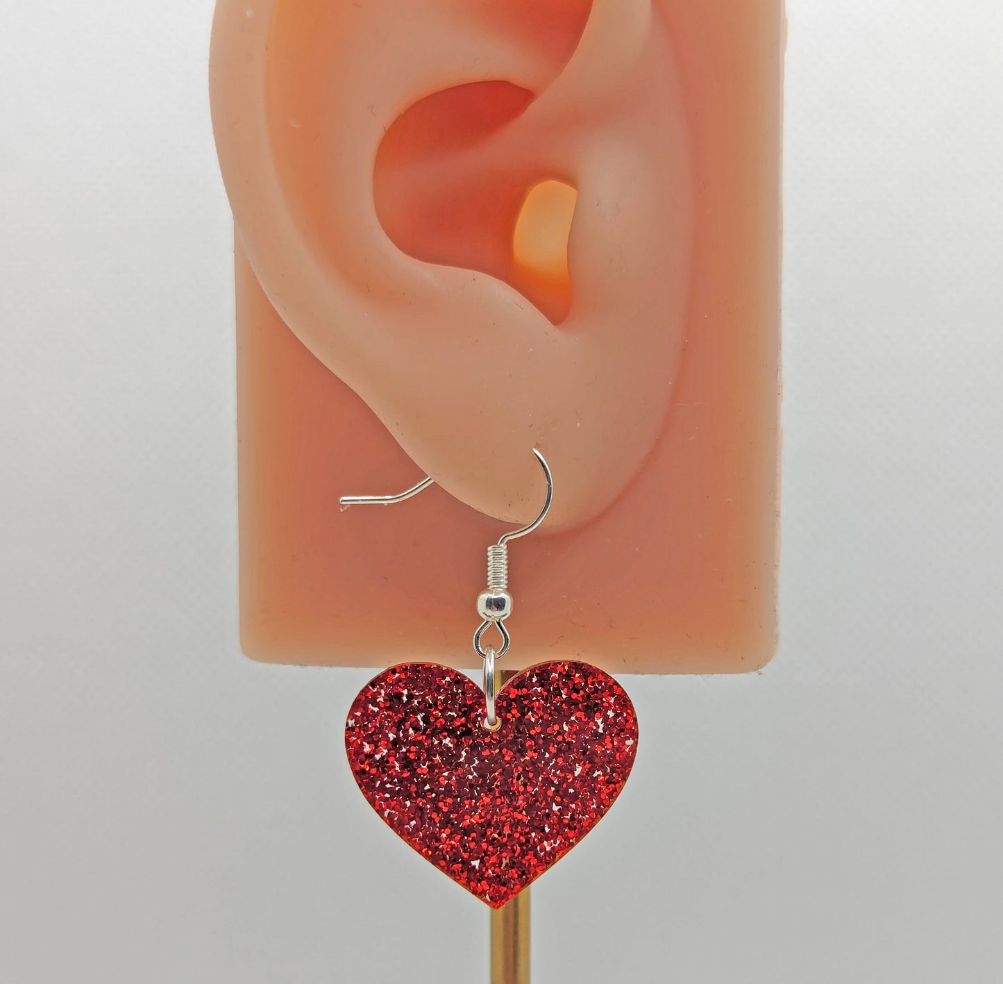 Image of a heart shaped earring on an ear showing the size against a white background. It is Red glitter acrylic and has silver fixings.