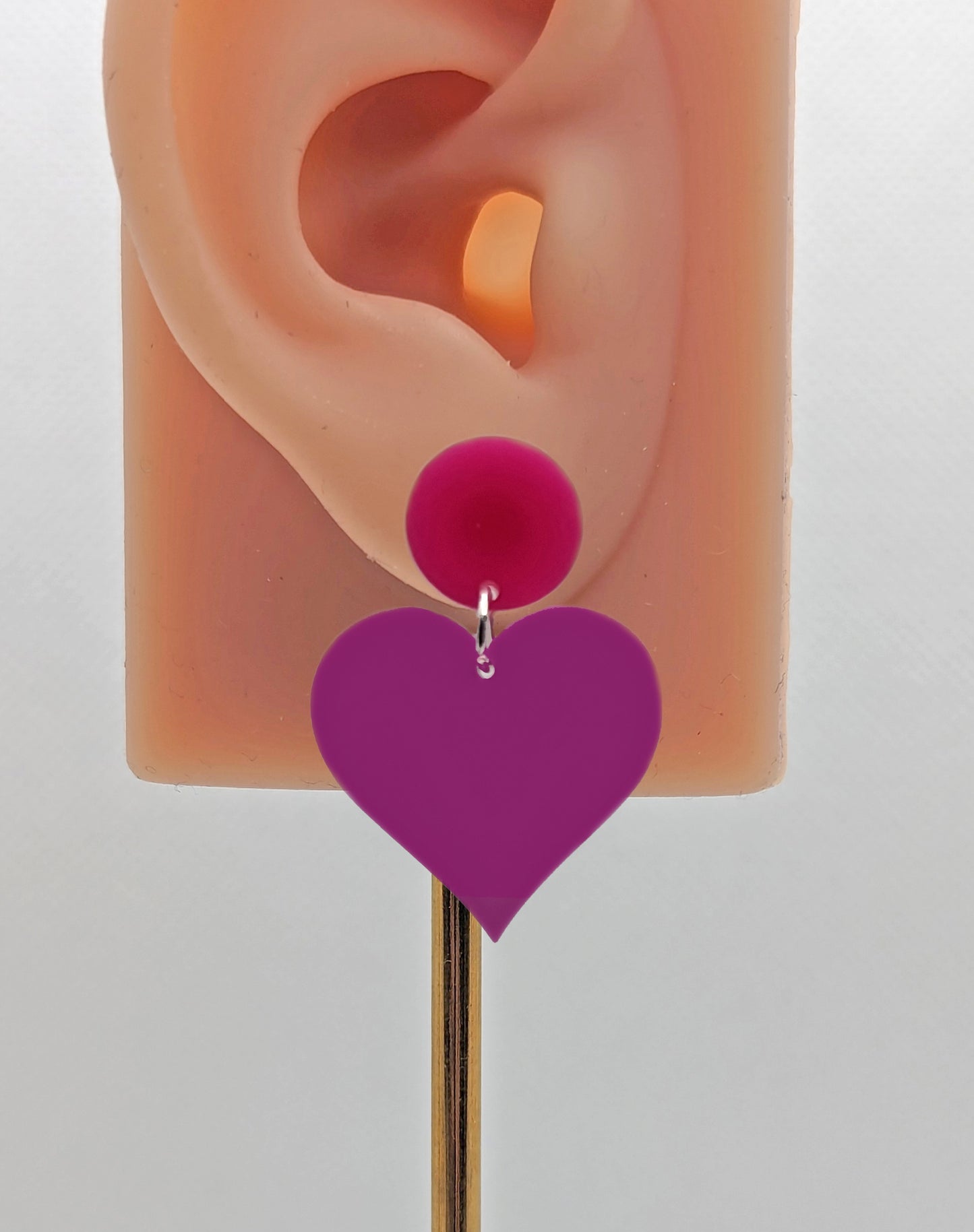 An earring with stud fixings on an ear to show the size. It is an acrylic circle, with a heart dangling underneath. The colour is a hot pink circle with Grape heart dangling underneath.