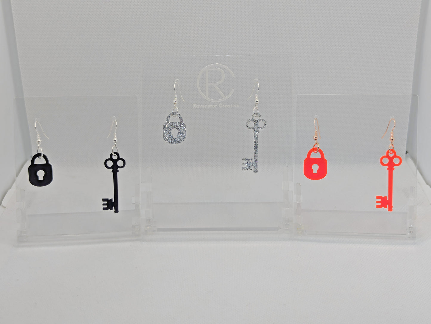 Three pairs of Two part drop earrings with ear wire hook fixings. Each set is a Lock and a Key. The colours are Black, Silver Glitter and Neon Pink. They are on clear stands with the Ravenstor Creative logo against a white background.