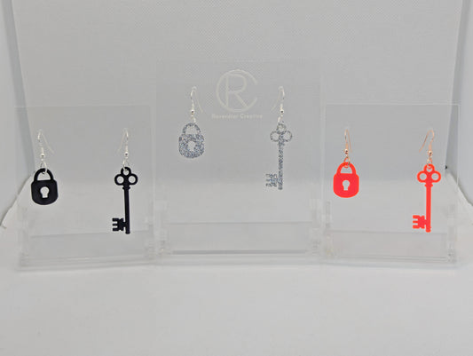 Three pairs of Two part drop earrings with ear wire hook fixings. Each set is a Lock and a Key. The colours are Black, Silver Glitter and Neon Pink. They are on clear stands with the Ravenstor Creative logo against a white background.