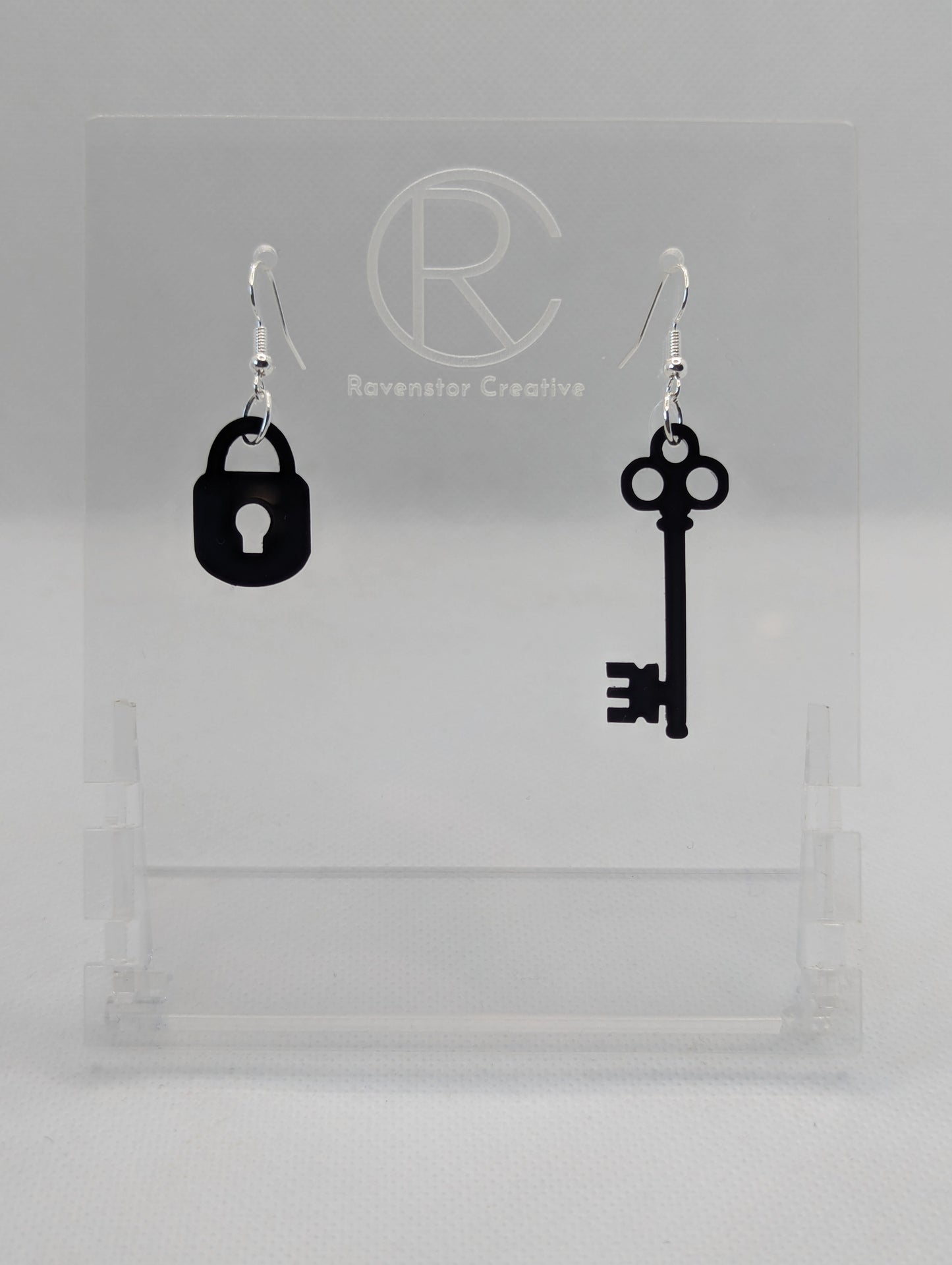 A set of two part drop earrings with Silver ear wire hook fixings. One earring is a Lock and the other is a Key. The colour for both is Black. They are on a clear stand with the Ravenstor Creative logo against a white background.