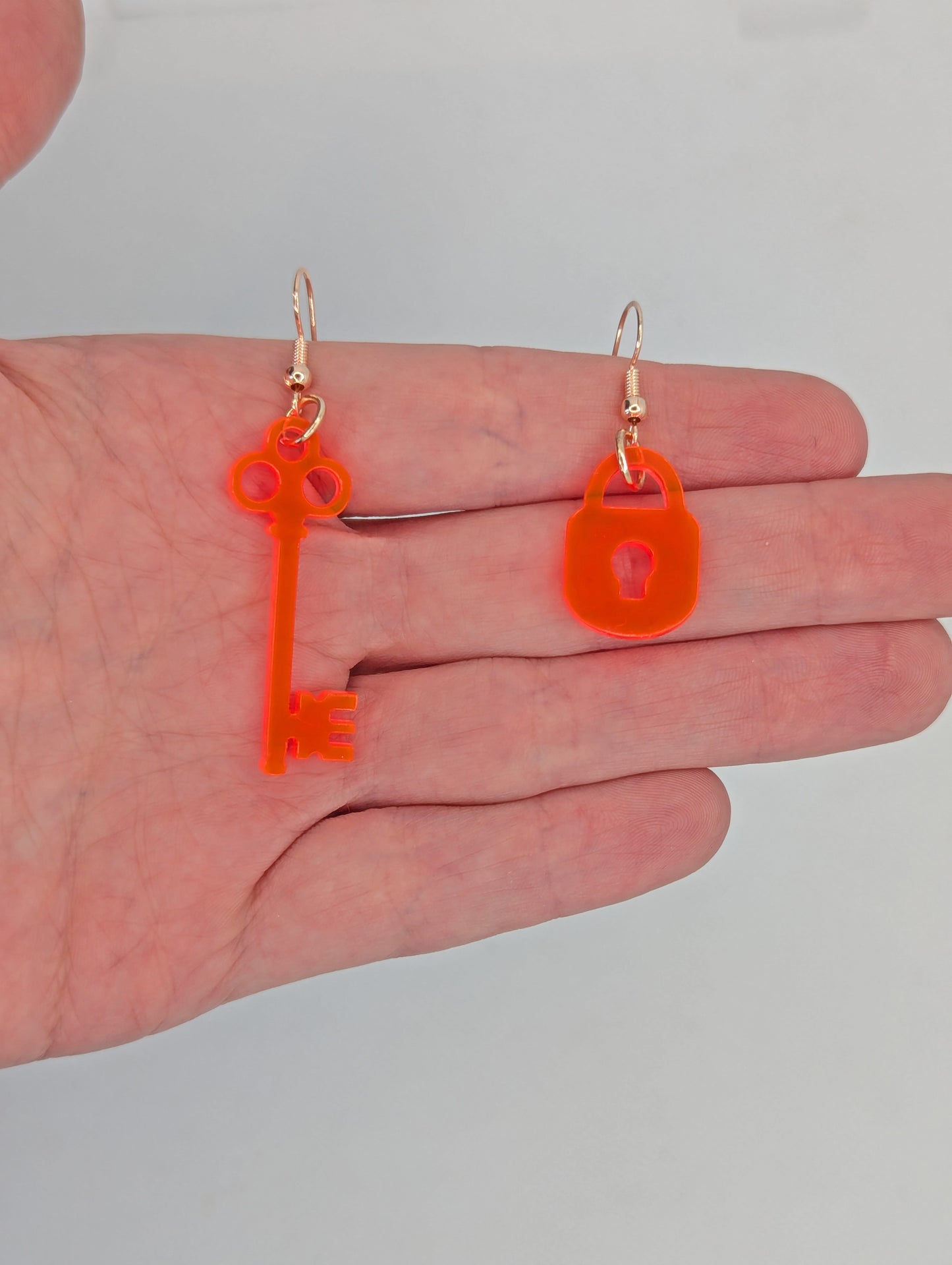 A set of two part drop earrings with rose gold ear wire hook fixings. One earring is a Lock and the other is a Key. They are against a hand to show the sizes. The key is two finger widths long and one wide. The Lock is one finger width long and wide.