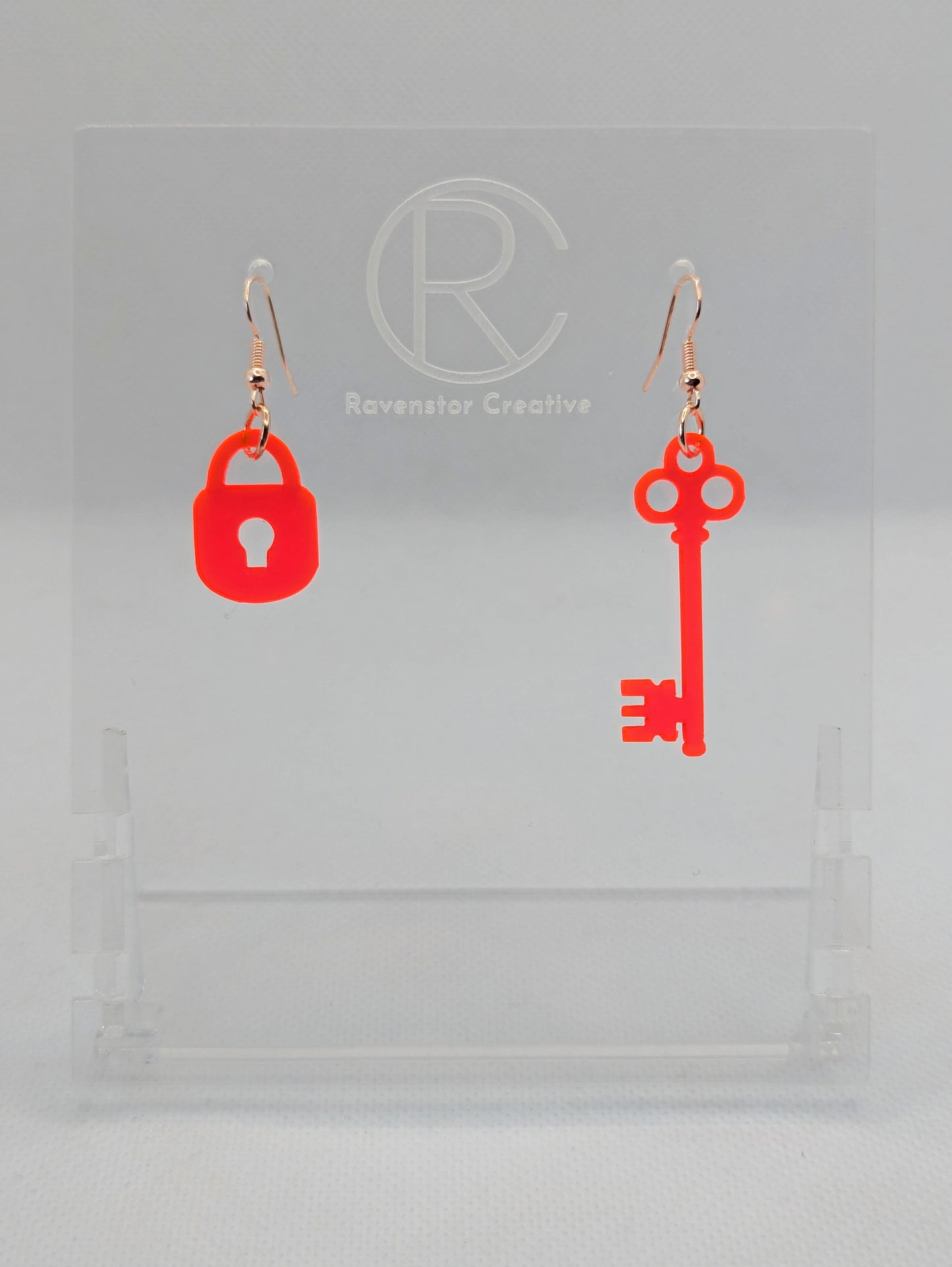 A set of two part drop earrings with Rose Gold ear wire hook fixings. One earring is a Lock and the other is a Key. The colour for both is Neon pink. They are on a clear stand with the Ravenstor Creative logo against a white background.