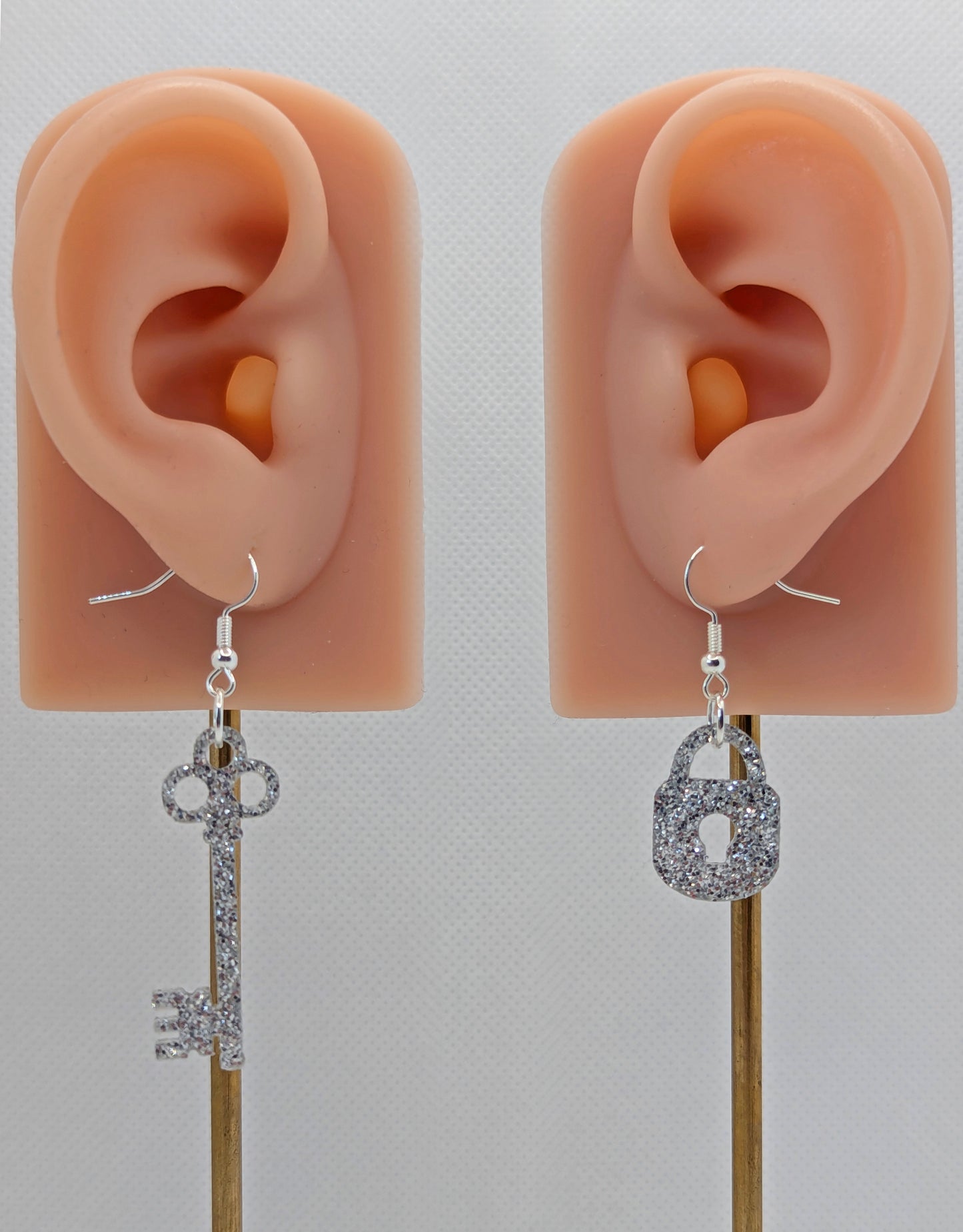 A set of two part drop earrings with Silver ear wire hook fixings. One earring is a Lock and the other is a Key. The colour for both is Silver Glitter. They are on ears to show the size of each one.