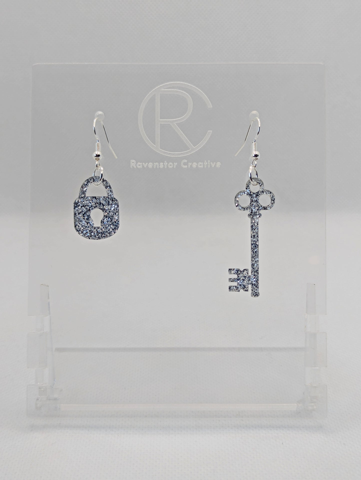A set of two part drop earrings with Silver ear wire hook fixings. One earring is a Lock and the other is a Key. The colour for both is Silver Glitter. They are on a clear stand with the Ravenstor Creative logo against a white background.