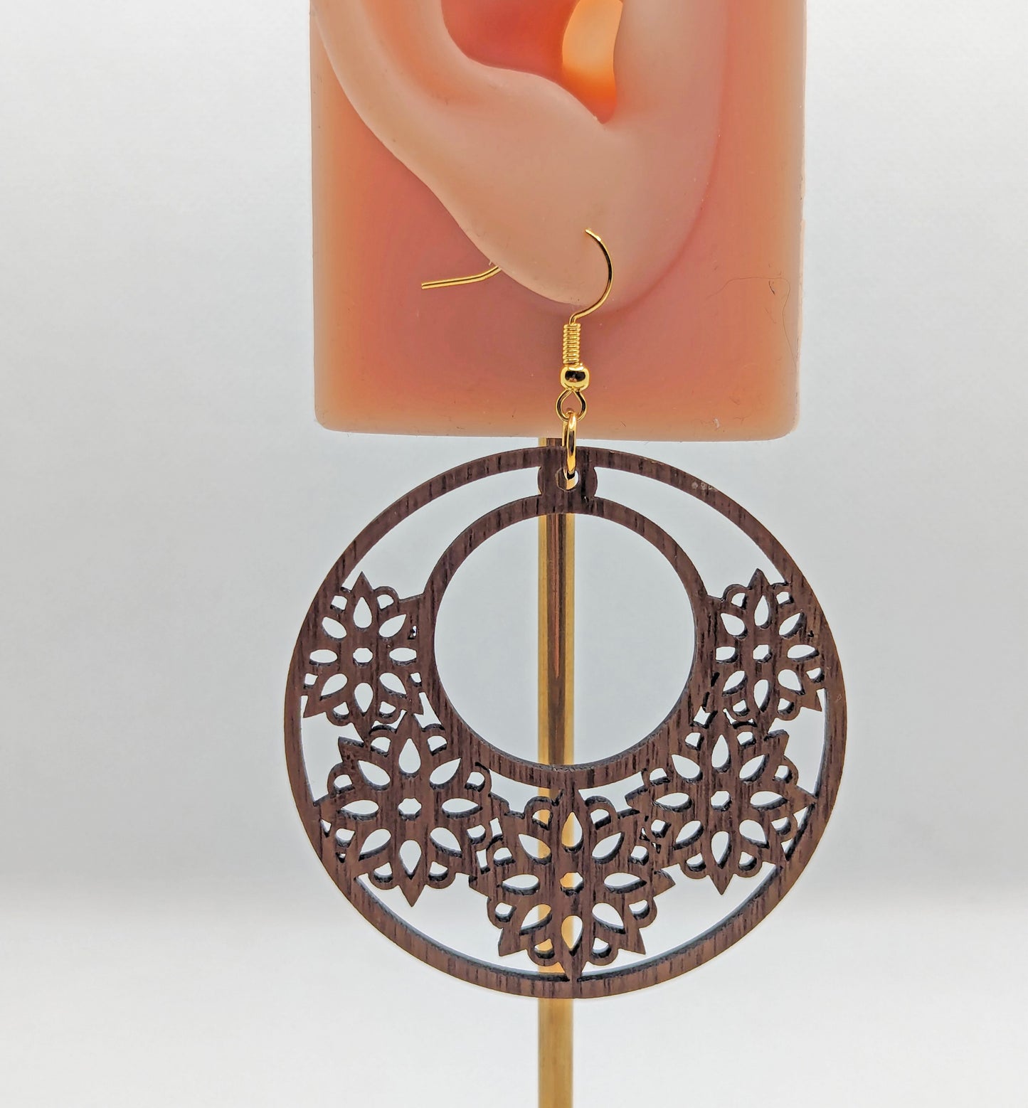 An wooden earring against an ear showing the size. It is a large hoop earring with flower / floral patterns cut in it with gold ear wire hook fixings. The colour is walnut wood.