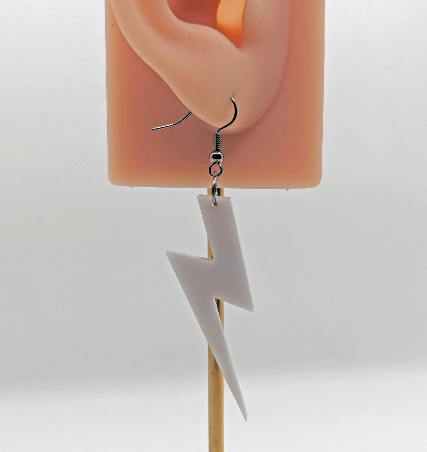 Image of a lightning bolt shaped earrings on an ear showing the size against a white background. They are made from grey acrylic and have black ear wire hook fixings.