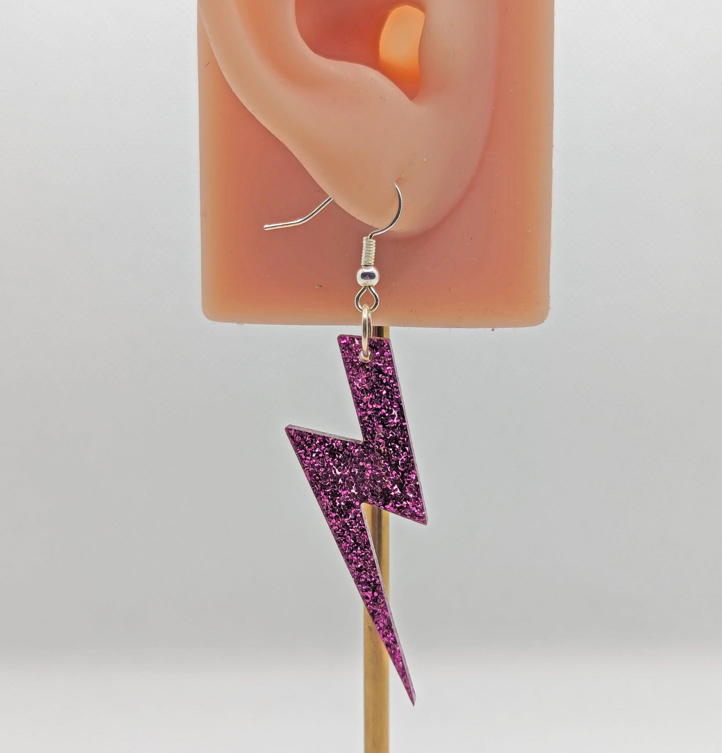 Image of a lightning bolt shaped earrings on an ear showing the size against a white background. They are made from Pink glitter acrylic and have silver ear wire hook fixings.