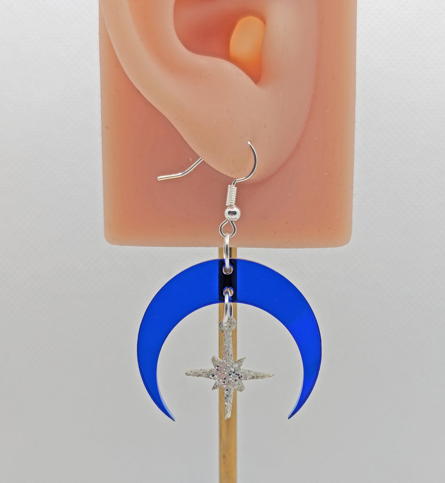 An earring that is two parts. silver ear wire fixings are attached to a crescent moon shaped frame with a small star dangling in the middle. They are on an ear showing the size against a white background.