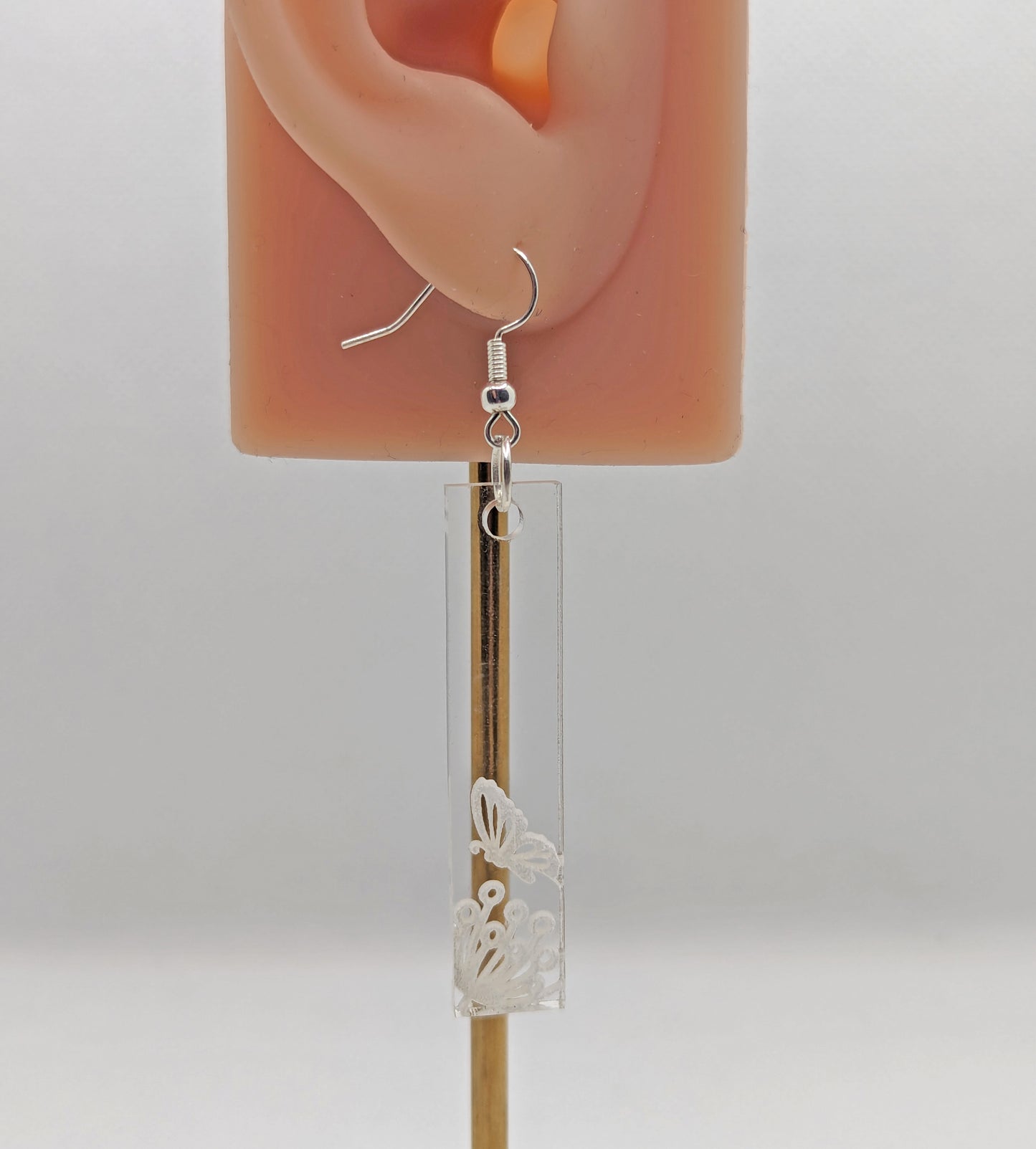 Image of a long slim rectangle earring with silver fixings on an ear showing the size. They are clear/see through and are engraved with a butterfly landing on a flower against a white background.