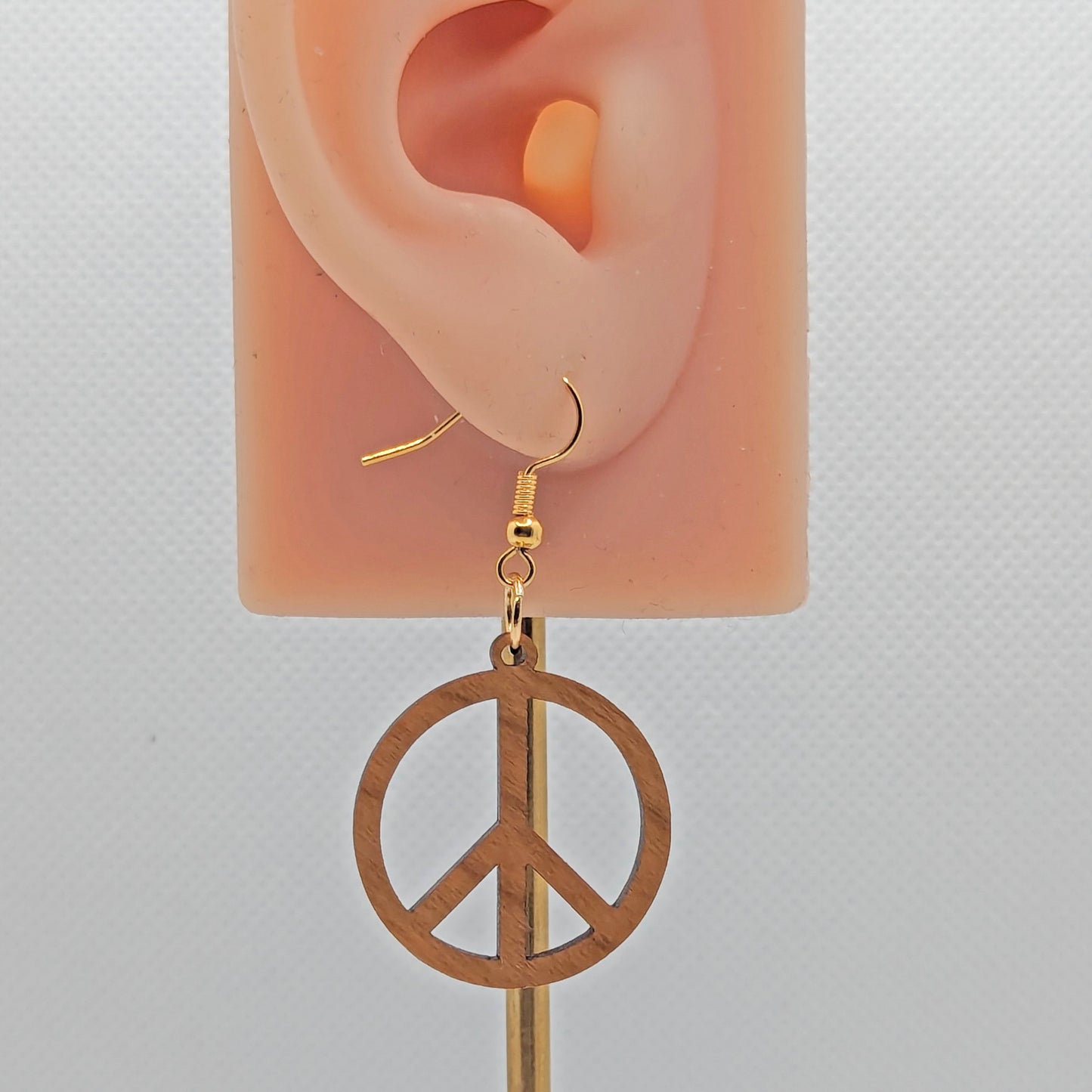 An earring in the shape of the 80s Retro Peace and love symbol. It has gold ear wire hook fixings and is made from Cherry wood. They are on an ear to show the size against a white background.