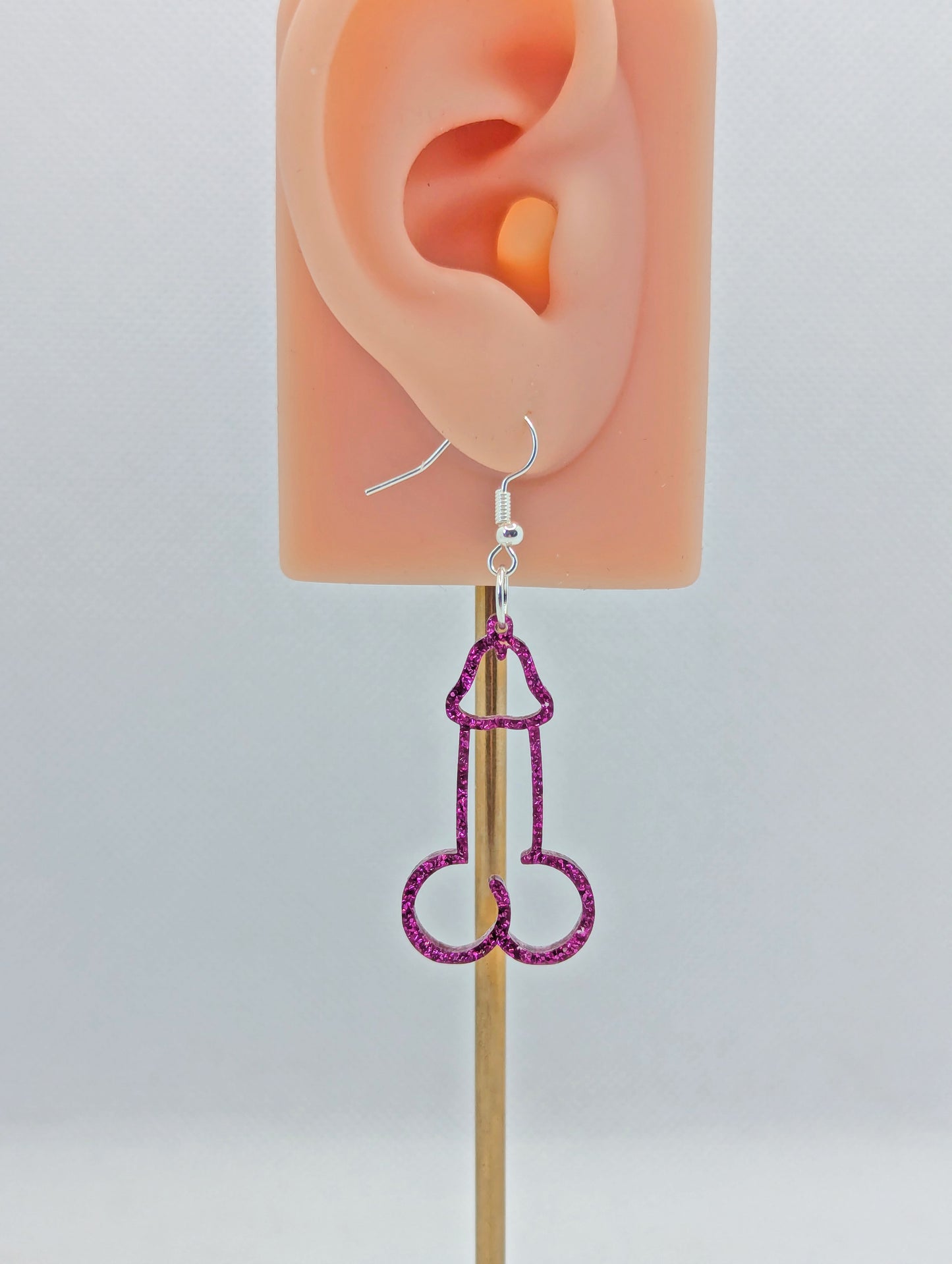 Image of an earring shaped like a Penis and Balls with a Silver ear wire hook fixing. These are ideal for funny Hen Dos or Bachelorette parties. The colour is Pink Glitter and is on an ear to show the size.