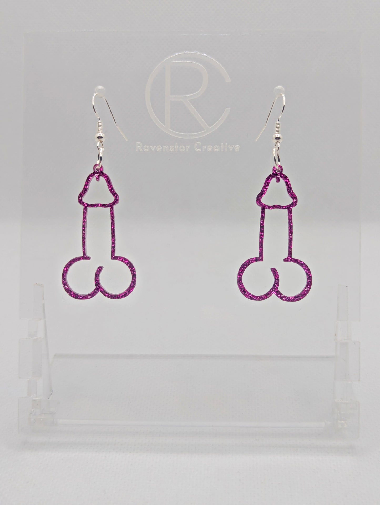 Image of a pair of earrings shaped like a Penis and Balls with Silver ear wire hook fixings. These are ideal for funny Hen Dos or Bachelorette parties. The colour is Pink Glitter and they are on clear stands against a white background.