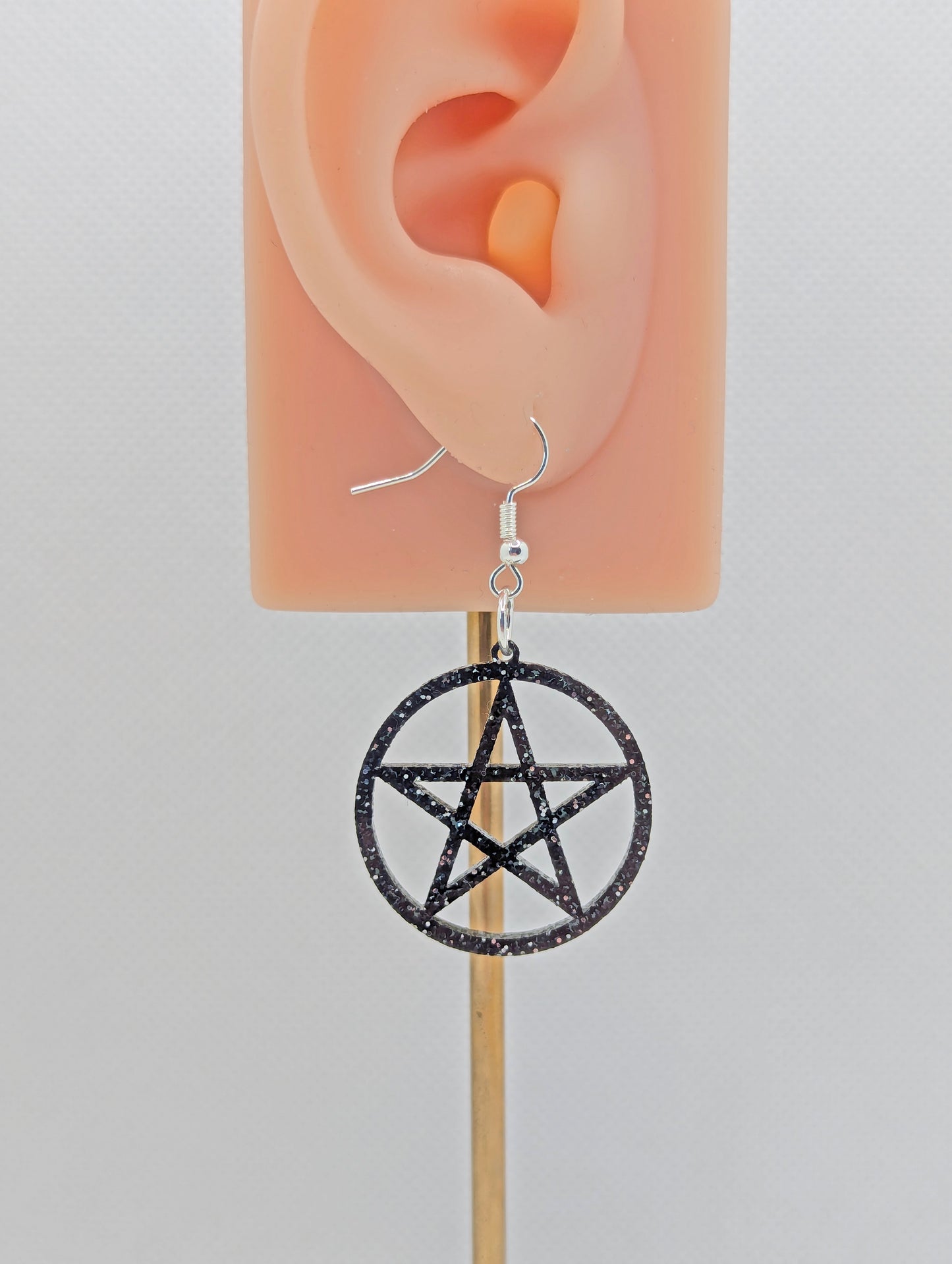 Image of a pair of Pentacle / Pentagram Symbol earrings on an ear showing the size against a white background. They are black and silver glitter and have black ear wire hook fixings.