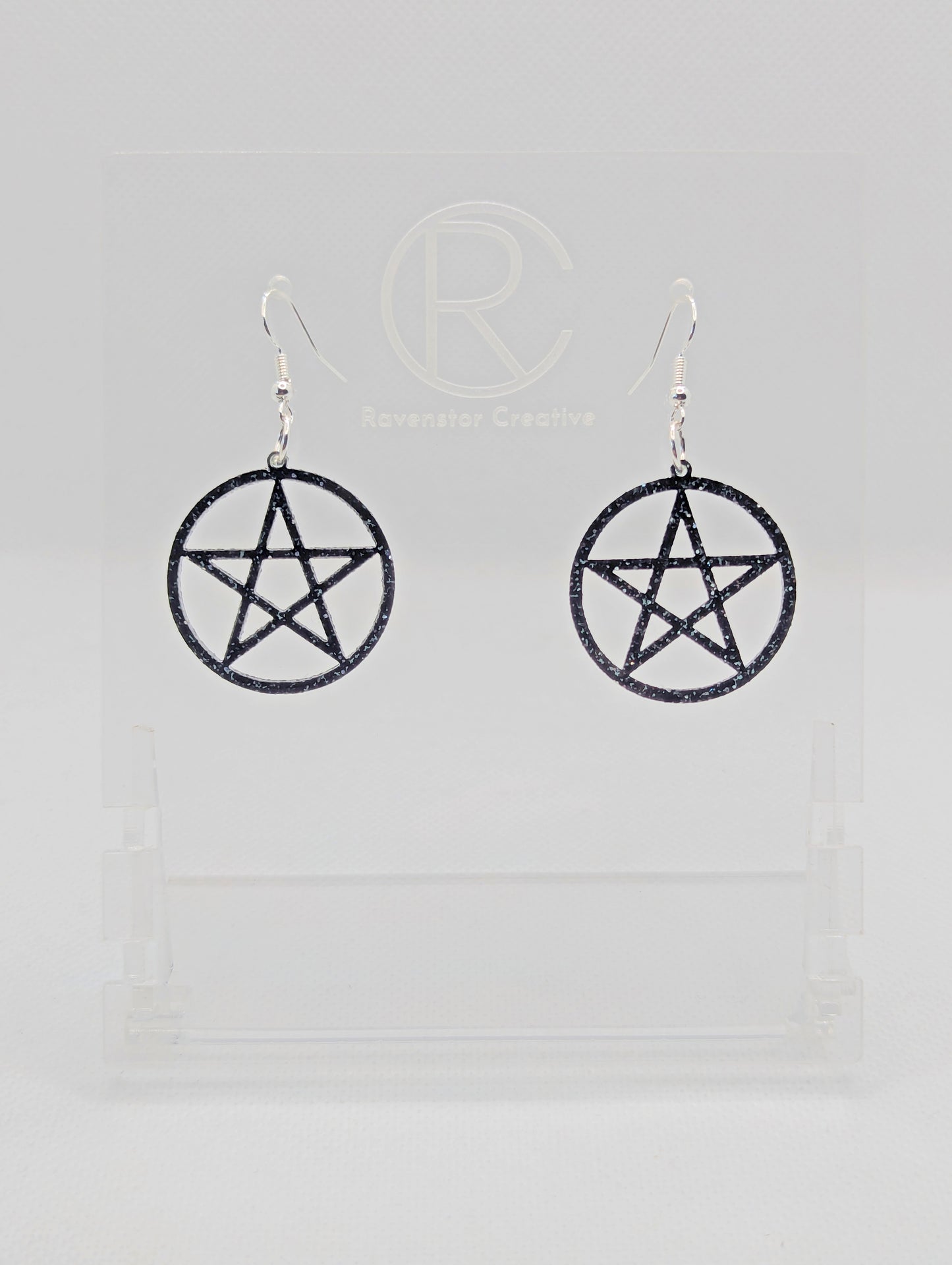 Shows a pair of earrings with Silver ear wire hook fixings. They are Pentacle / Pentagram Symbols. The colour of them are Black and Silver Glitter acrylic. They are on a see through stand that has the Ravenstor Creative Logo and its against a white background