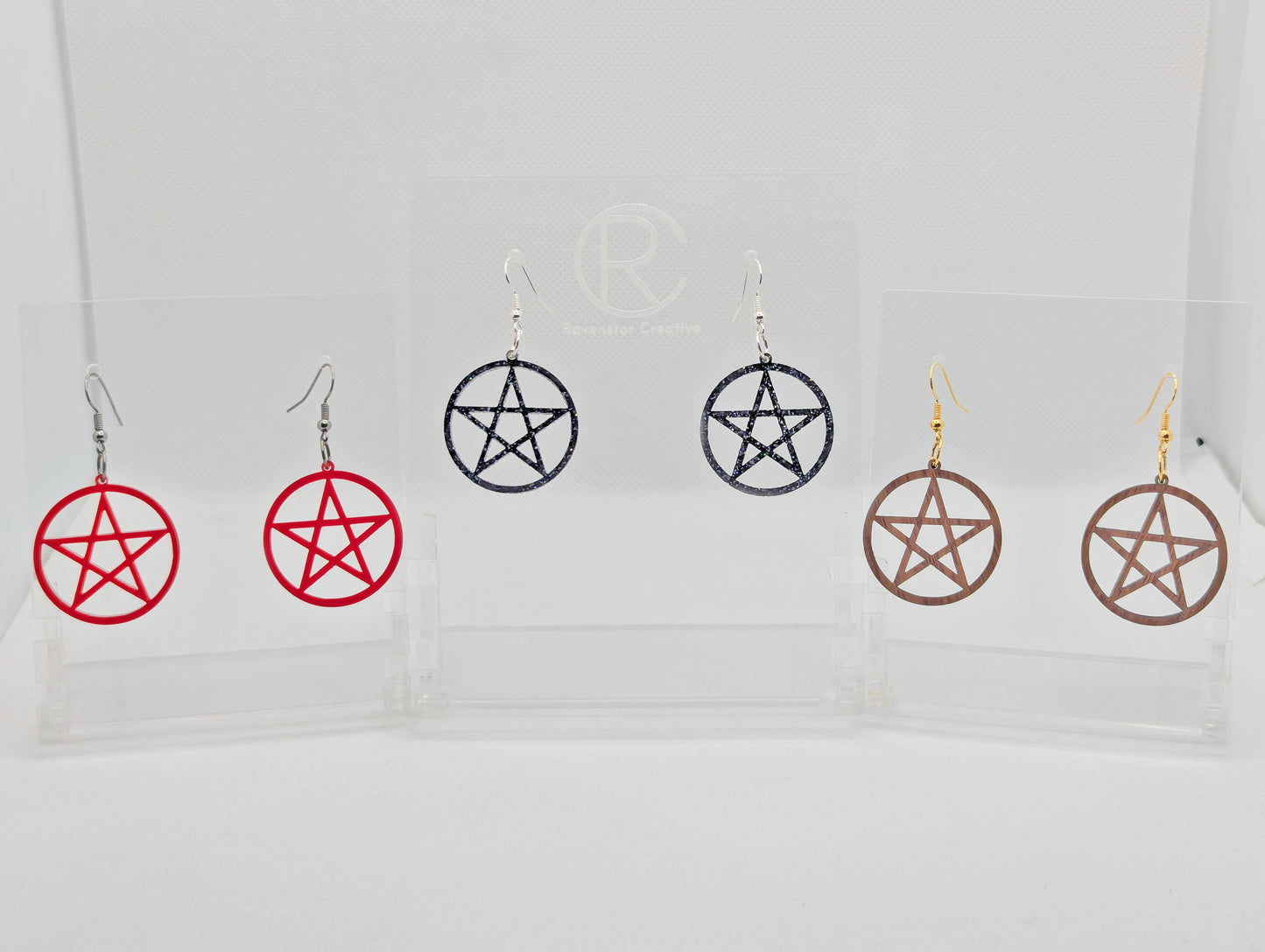 Shows three pairs of earrings with ear wire hook fixings. They are Pentacle / Pentagram symbols. The colours of them are Translucent Red, Black and Silver Glitter and Walnut Wood. They are against a white background on clear stands.