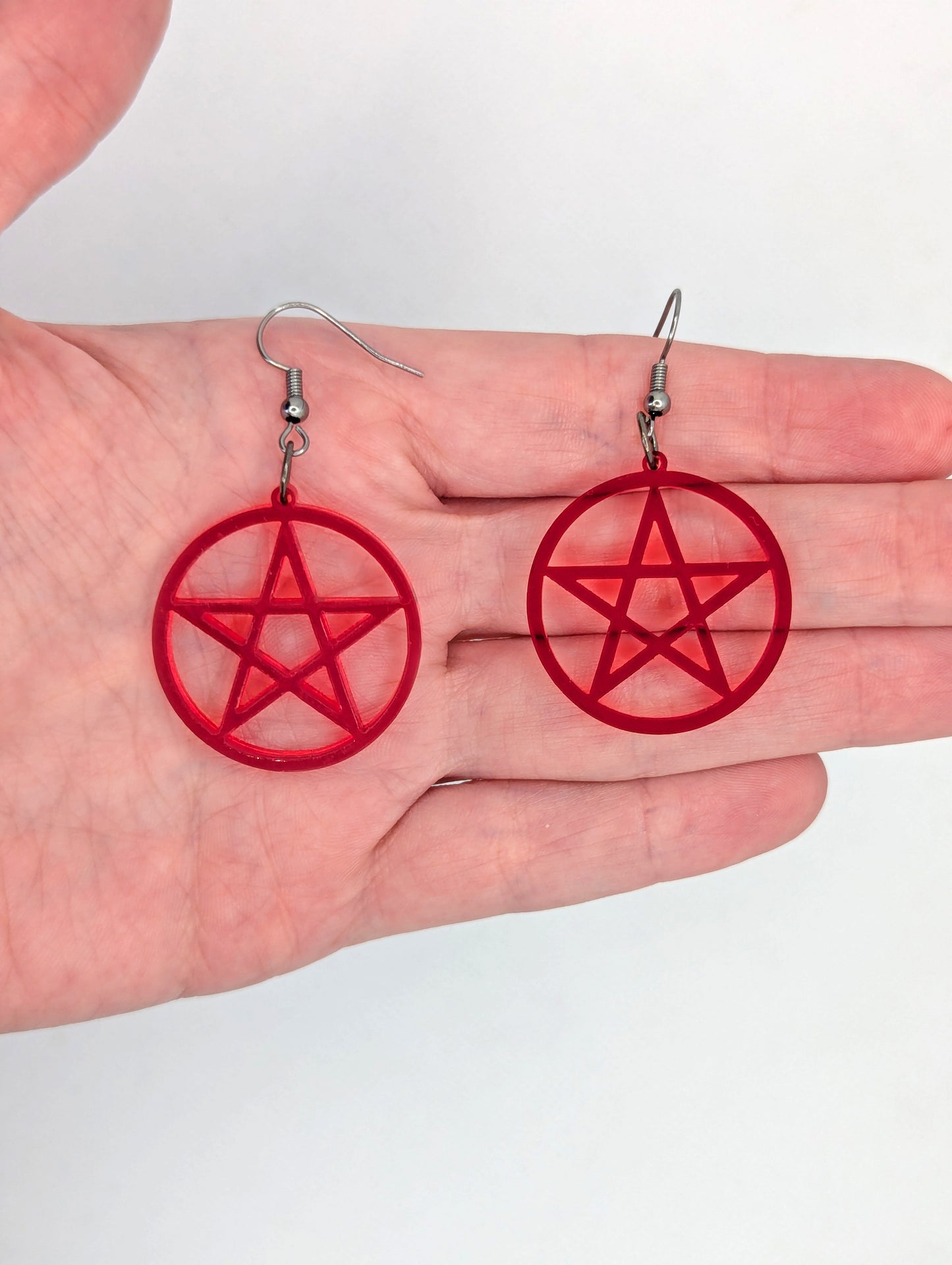 Shows a pair of earrings with black ear wire hook fixings. They are Pentacle / Pentagram Symbols. The colour of them are translucent red acrylic. They are against a hand to show the size of two finger widths length and wide.