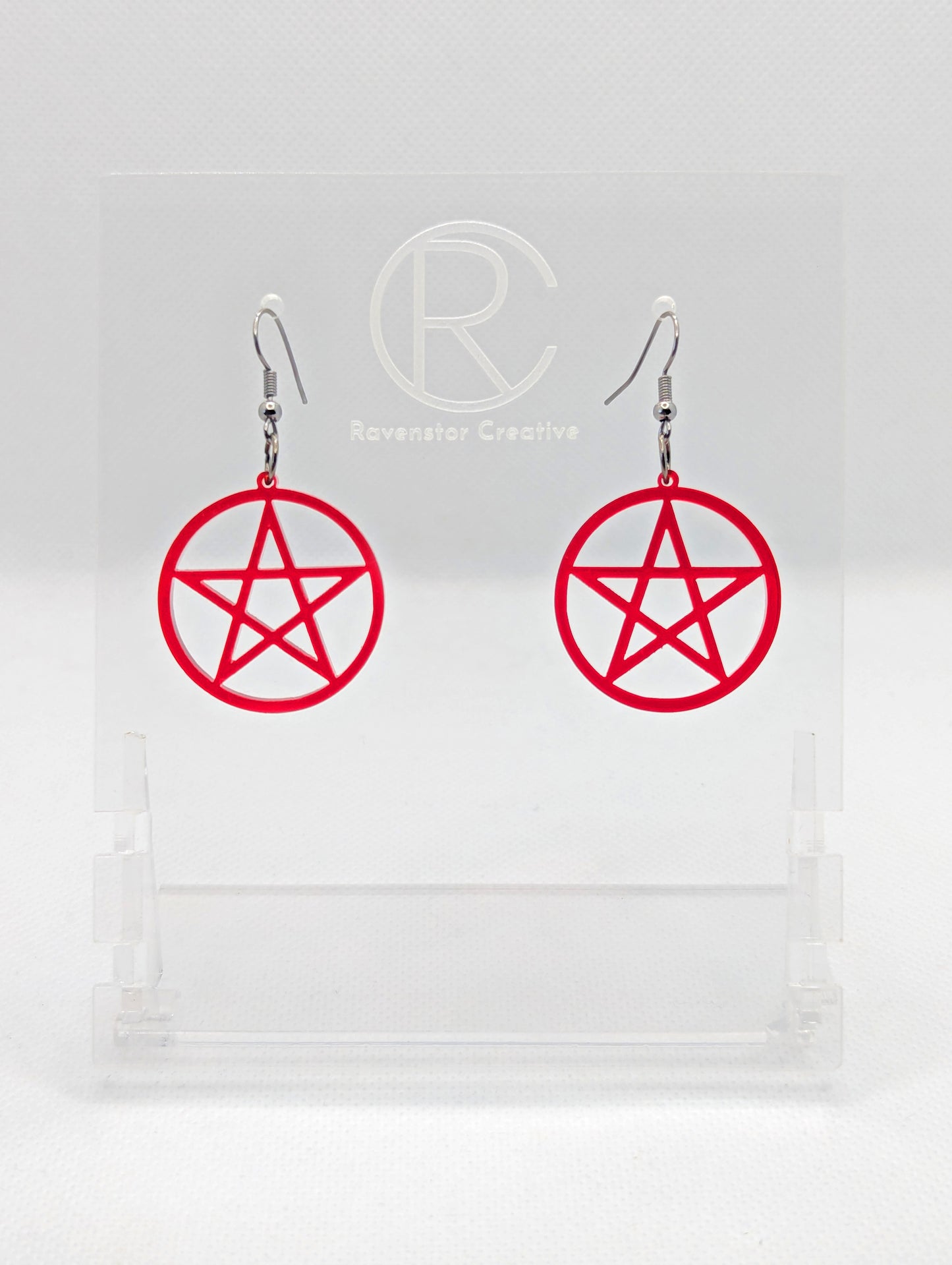 Shows a pair of earrings with black ear wire hook fixings. They are Pentacle / Pentagram Symbols. The colour of them are translucent red acrylic. They on a see through stand that has the Ravenstor Creative Logo and its against a white background